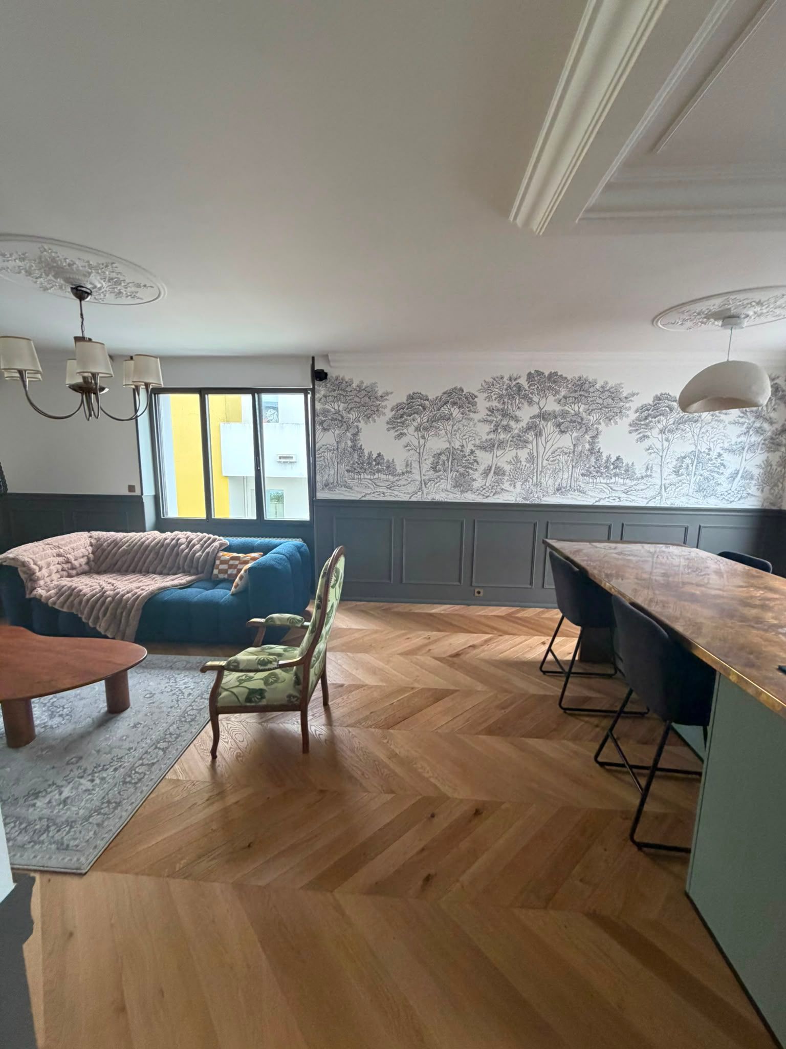 Salon moderne avec parquet à chevrons, canapé bleu, table à manger et fresque murale à motifs