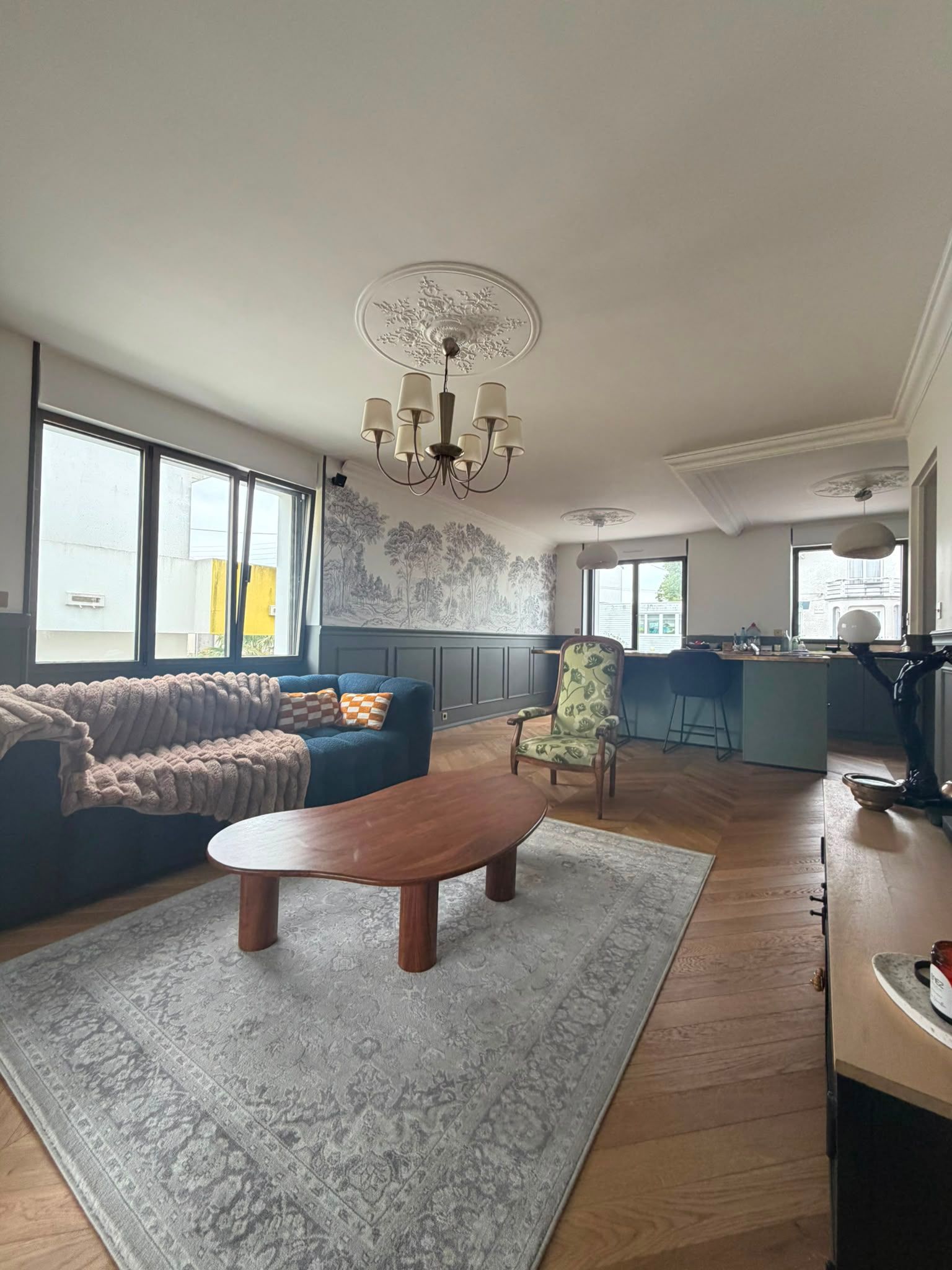 Salon lumineux avec des canapés bleus, une table basse en bois, un tapis gris, un lustre et de grandes fenêtres.