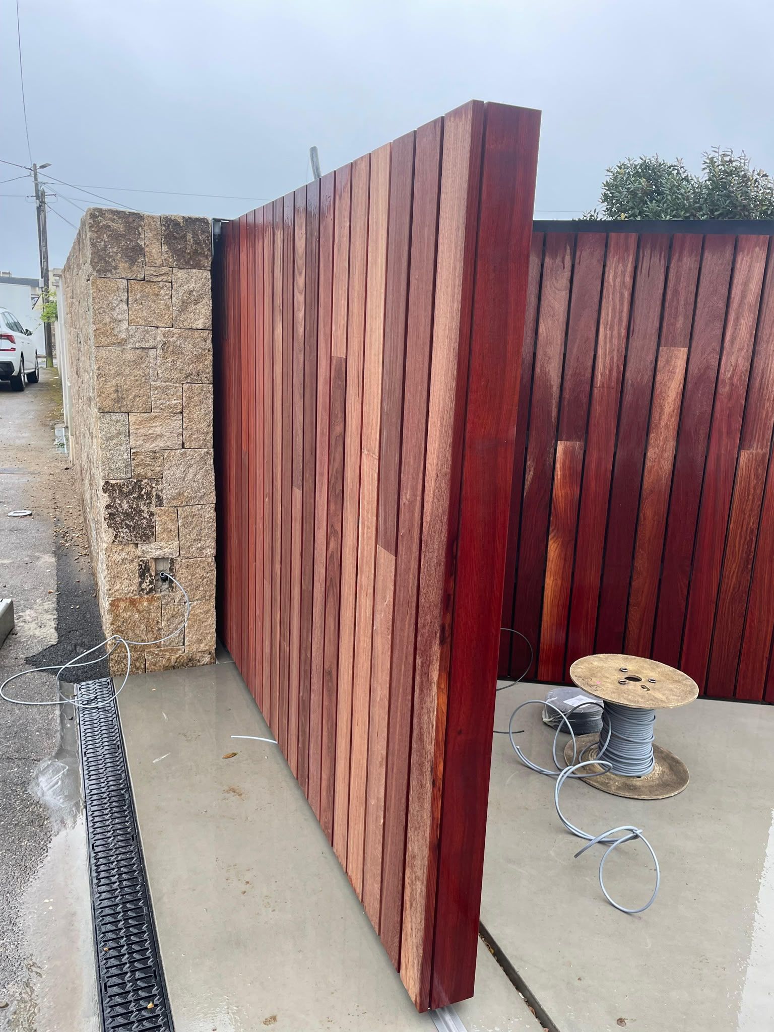 Panneaux de clôture en bois rouge le long d’un mur de pierre, avec une bobine de câble et des outils sur une allée en béton.