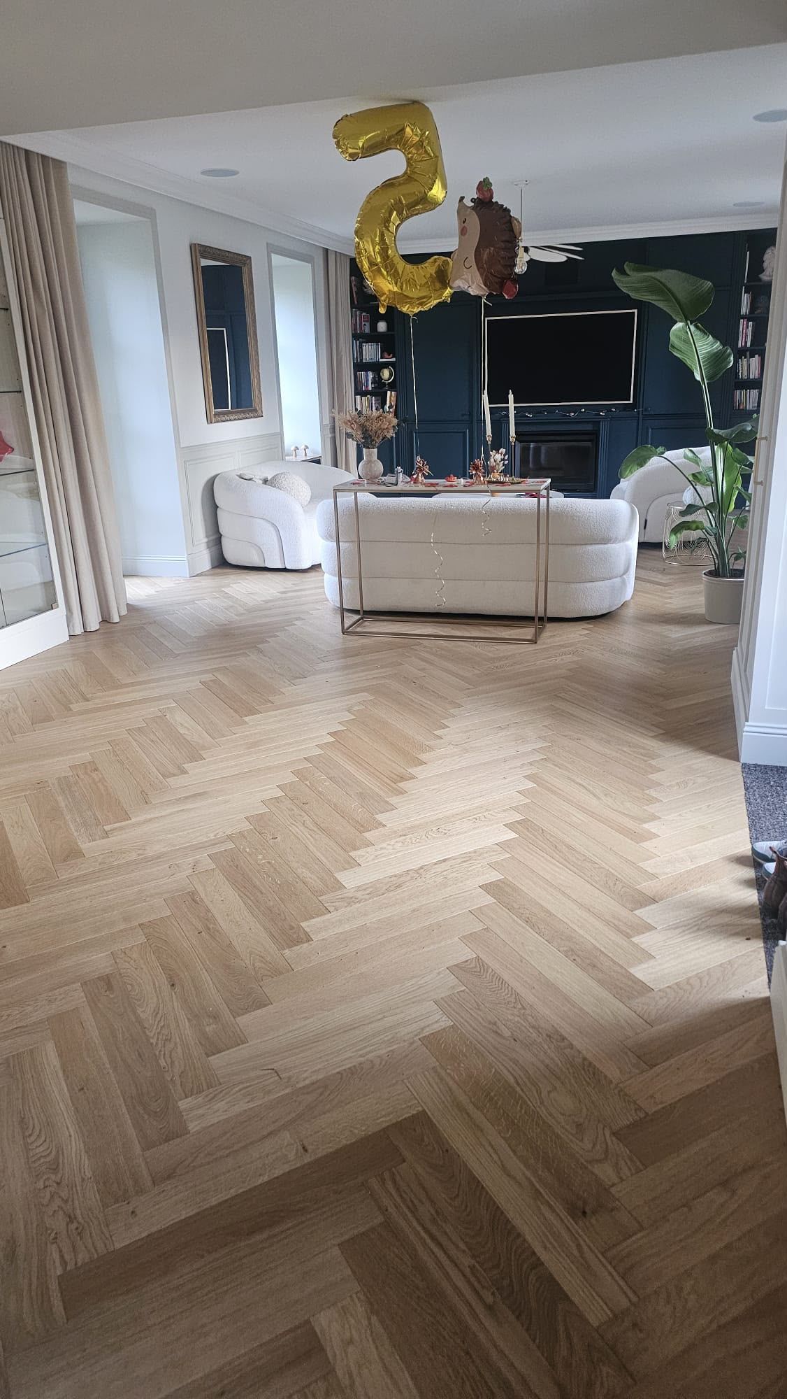 Salon avec parquet en chevrons, canapé blanc, mur TV noir et décorations avec des ballons dorés