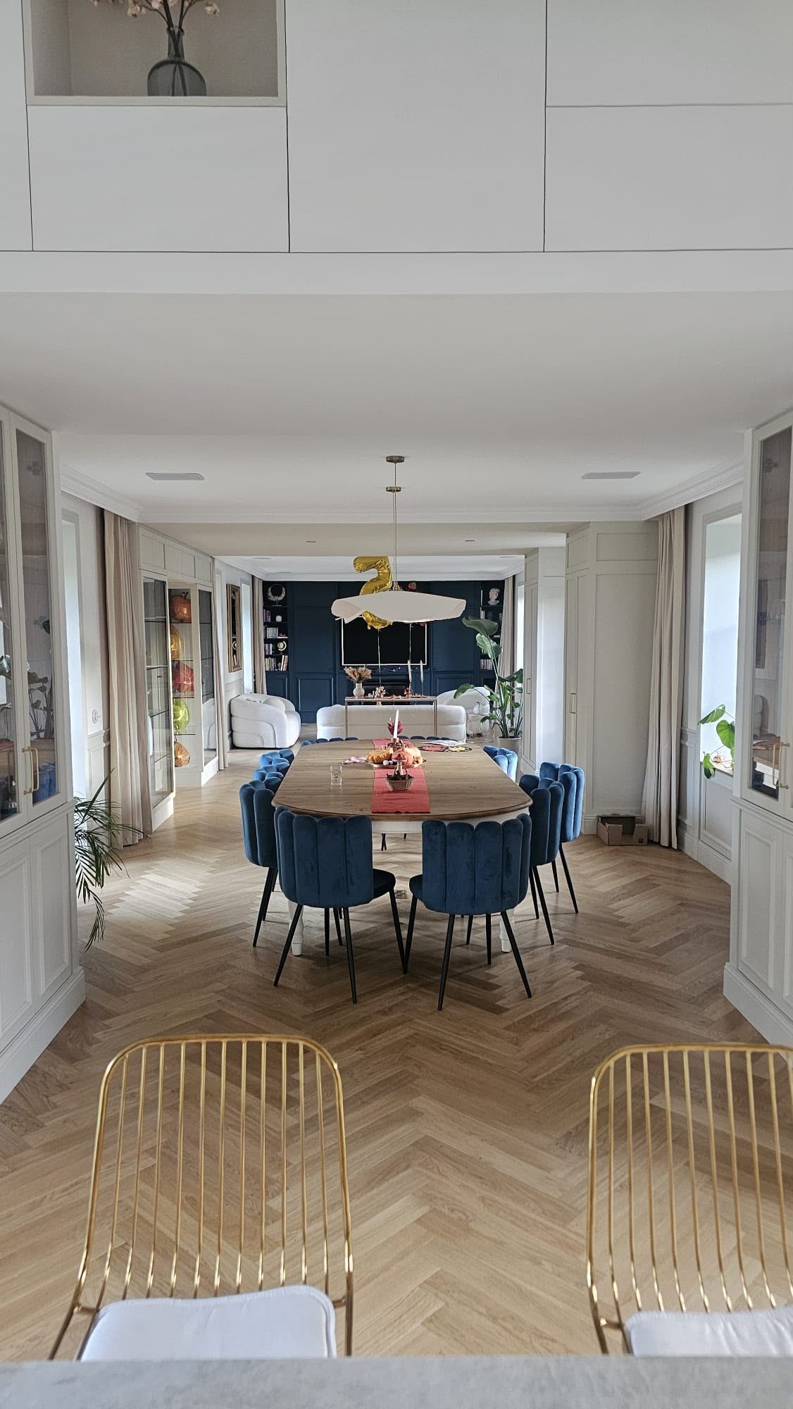 Salle à manger élégante avec une longue table, des chaises bleues et un lustre, vue depuis une porte ouverte.