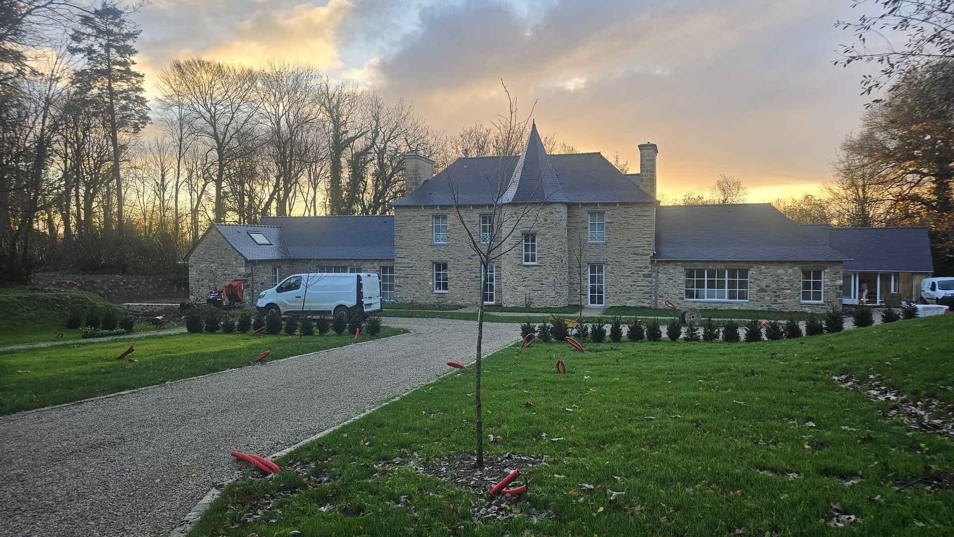 Manoir en pierre au coucher du soleil, avec une allée en gravier, une pelouse verdoyante et des arbres en arrière-plan
