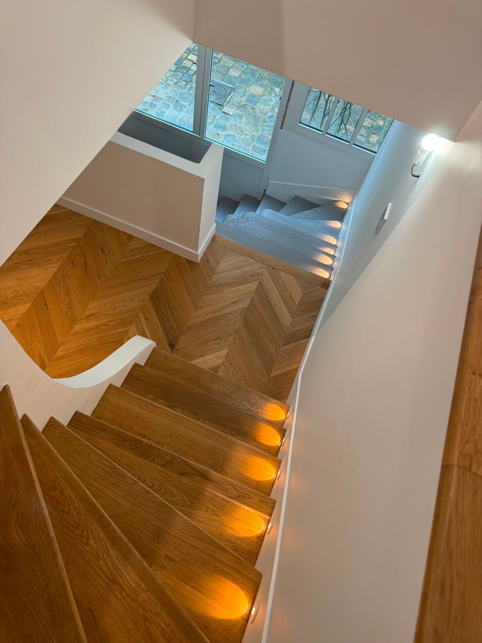 Escalier en bois dans une maison moderne, éclairé par une lumière chaleureuse, menant à un palier lumineux doté de fenêtres.