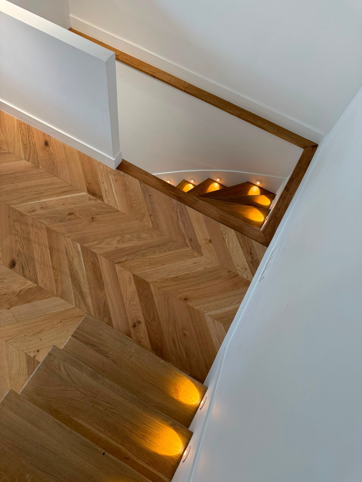 Escalier en bois avec parquet à chevrons et éclairage chaleureux intégré aux marches, le long d’une rampe blanche