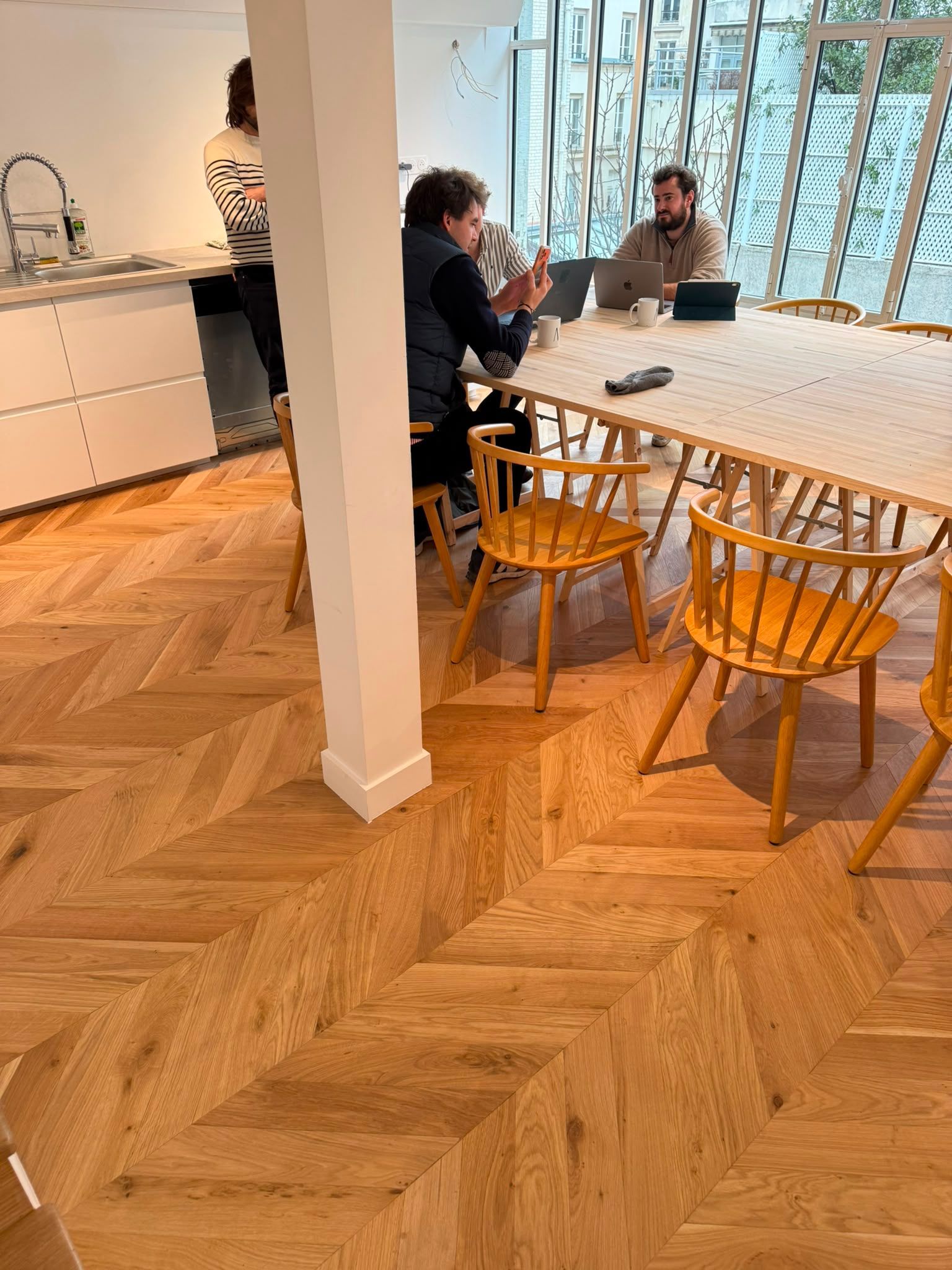 Deux personnes discutent autour d’une table dans une cuisine moderne et lumineuse, au sol en parquet à chevrons.