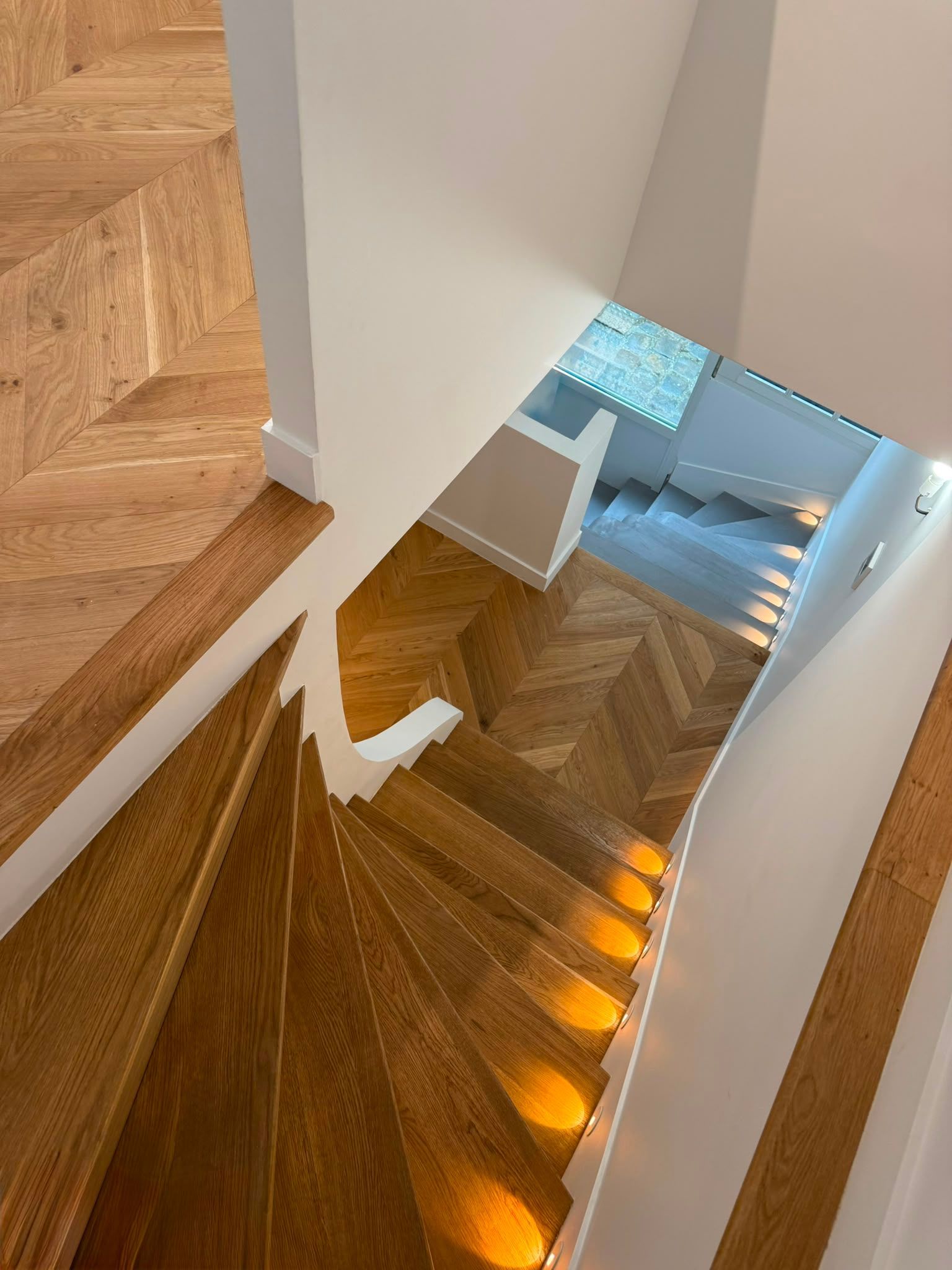 Vue plongeante sur un escalier en colimaçon en bois, avec des murs blancs et un palier éclairé par une fenêtre en contrebas.