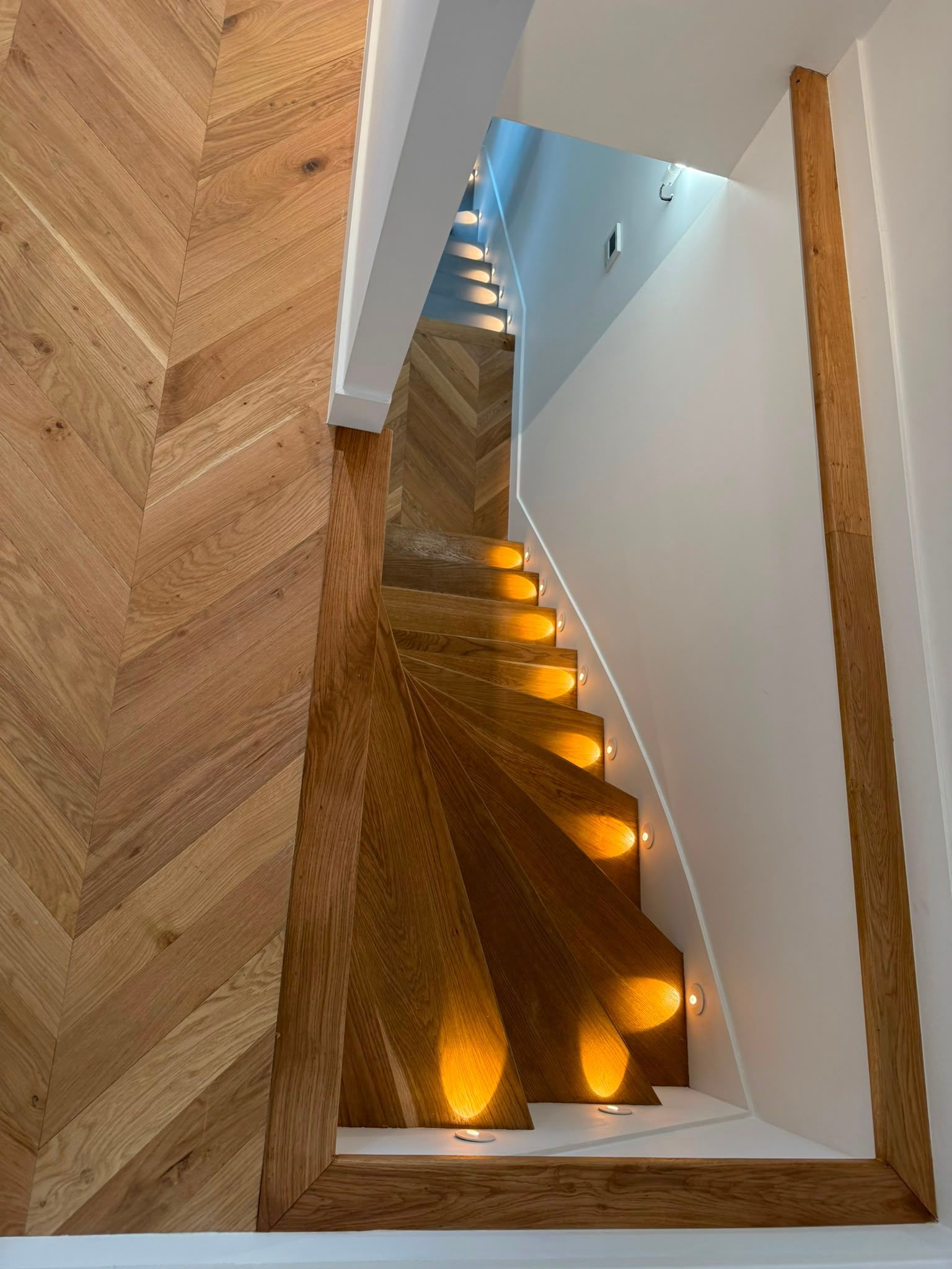 Escalier moderne en bois, éclairé par une lumière chaude intégrée aux marches, avec des murs blancs, vu depuis une porte