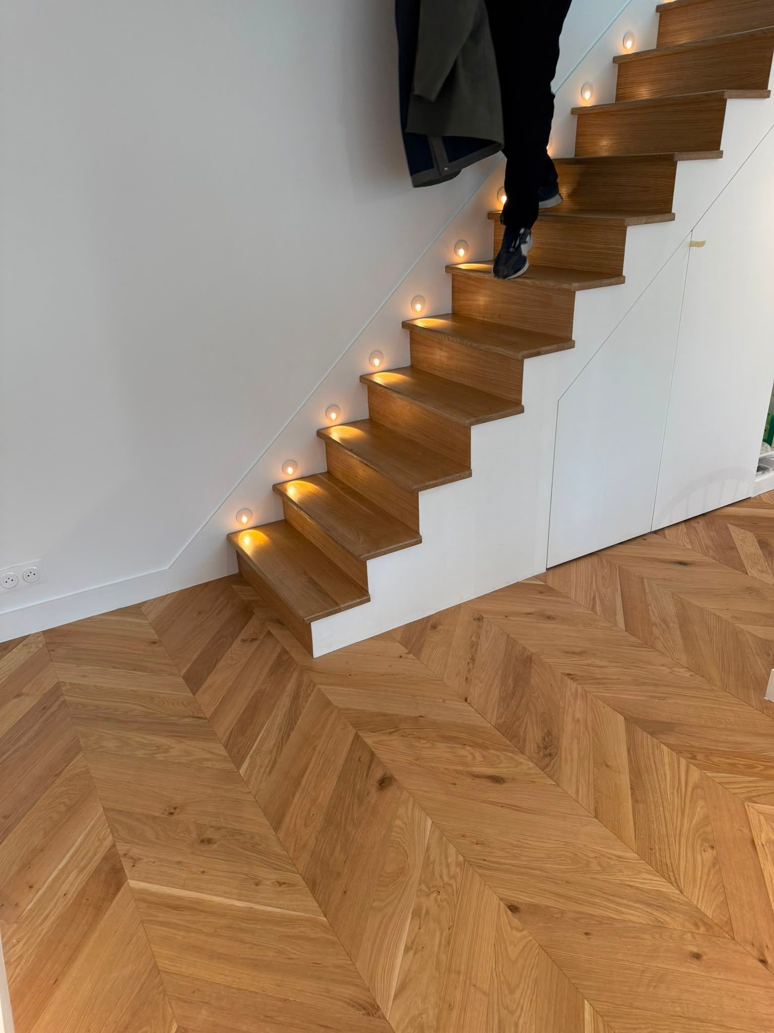 Escalier en bois orné de guirlandes lumineuses chaleureuses, près d’un parquet en chevrons et de murs blancs