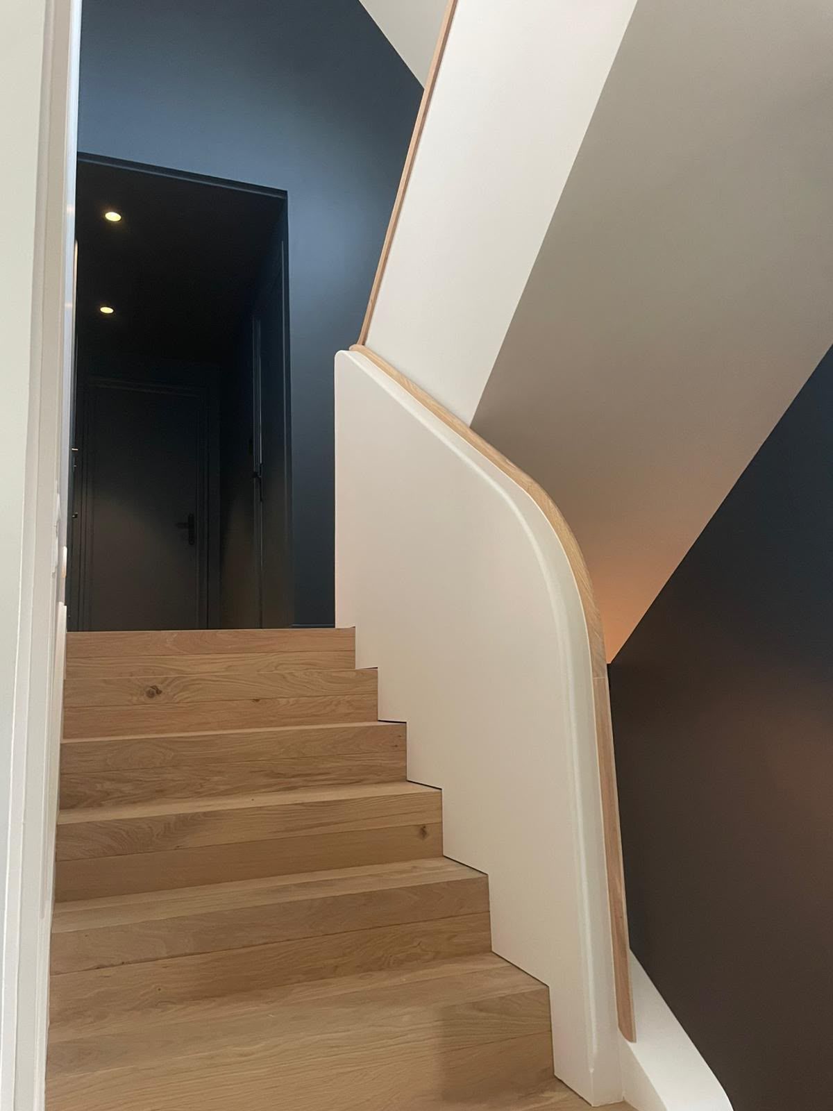 Escalier en bois menant à une porte sombre, avec des murs blancs et une rampe incurvée.