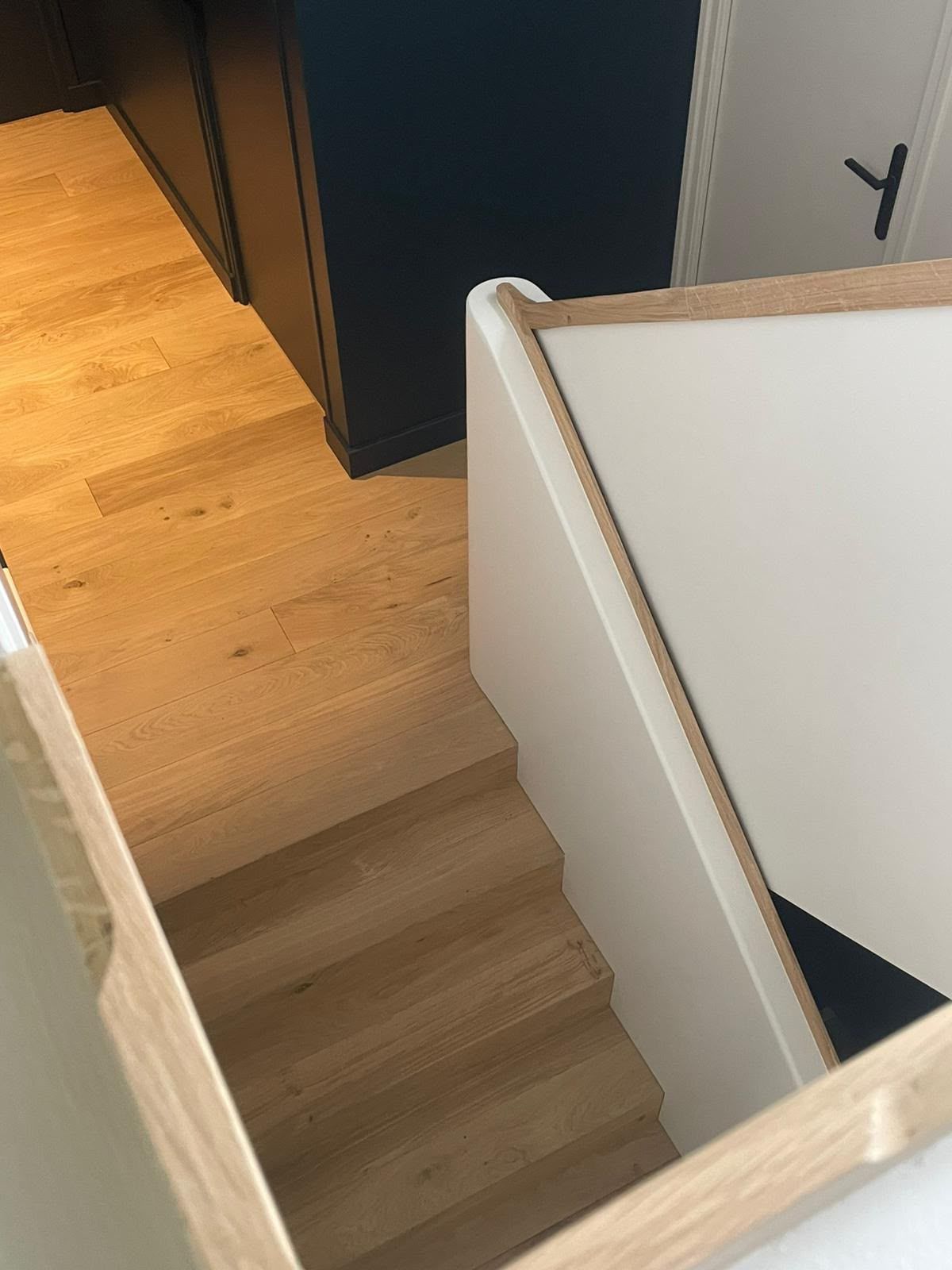 Cage d’escalier intérieure avec des marches en bois clair et des murs blancs, à côté de meubles sombres