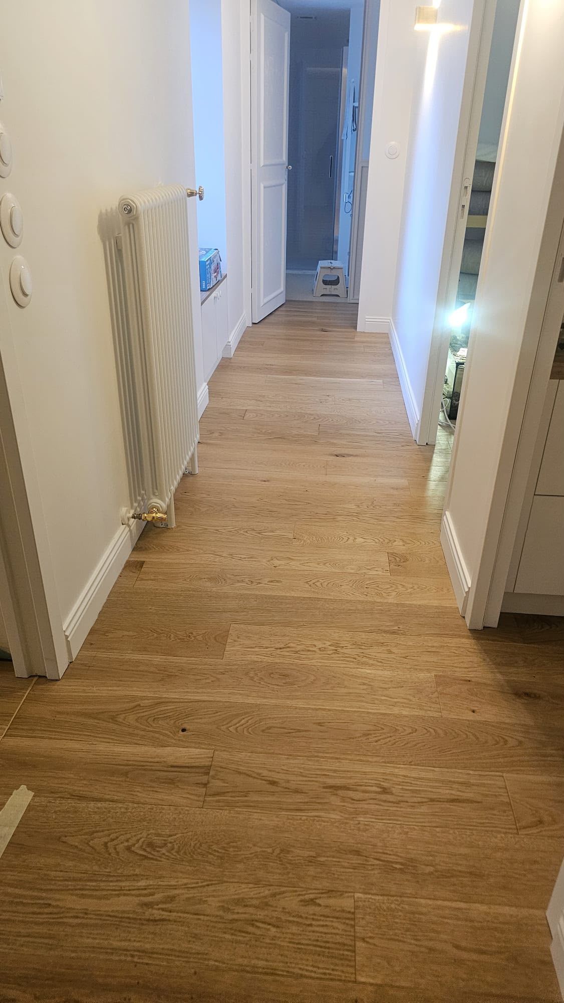 Couloir étroit avec un parquet clair, des murs blancs, un radiateur et une porte ouverte au fond