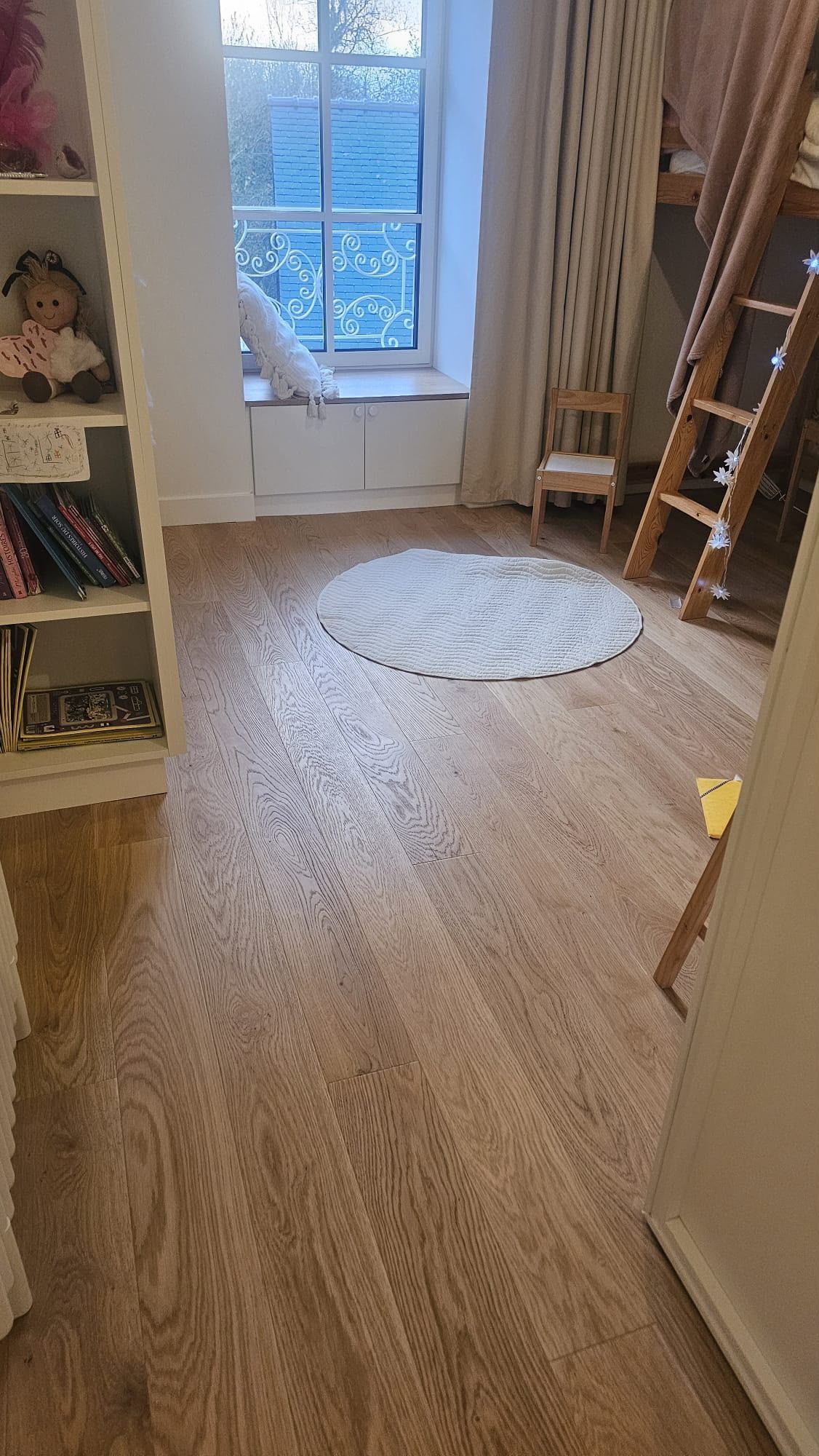 Petite chambre avec parquet, rideaux blancs, tapis rond et bibliothèque près de la fenêtre.
