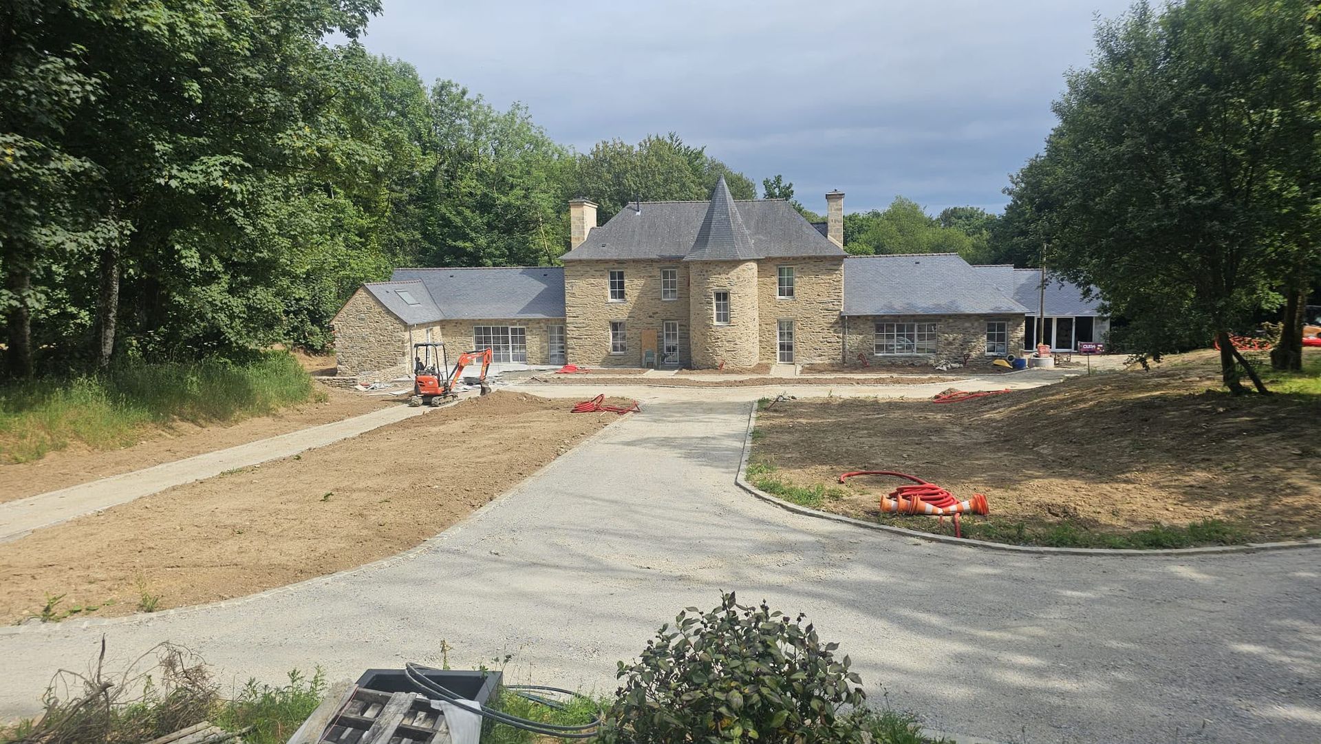 Maison en pierre en construction, avec une allée incurvée, une cour en terre et des ouvriers près de l’entrée