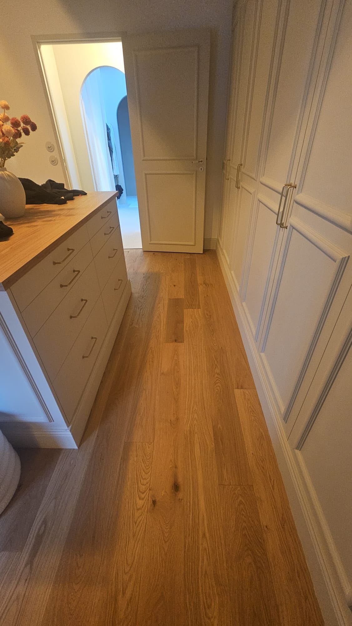 Couloir étroit avec un parquet, des murs blancs lambrissés et une commode à gauche, menant à une porte ouverte éclairée.