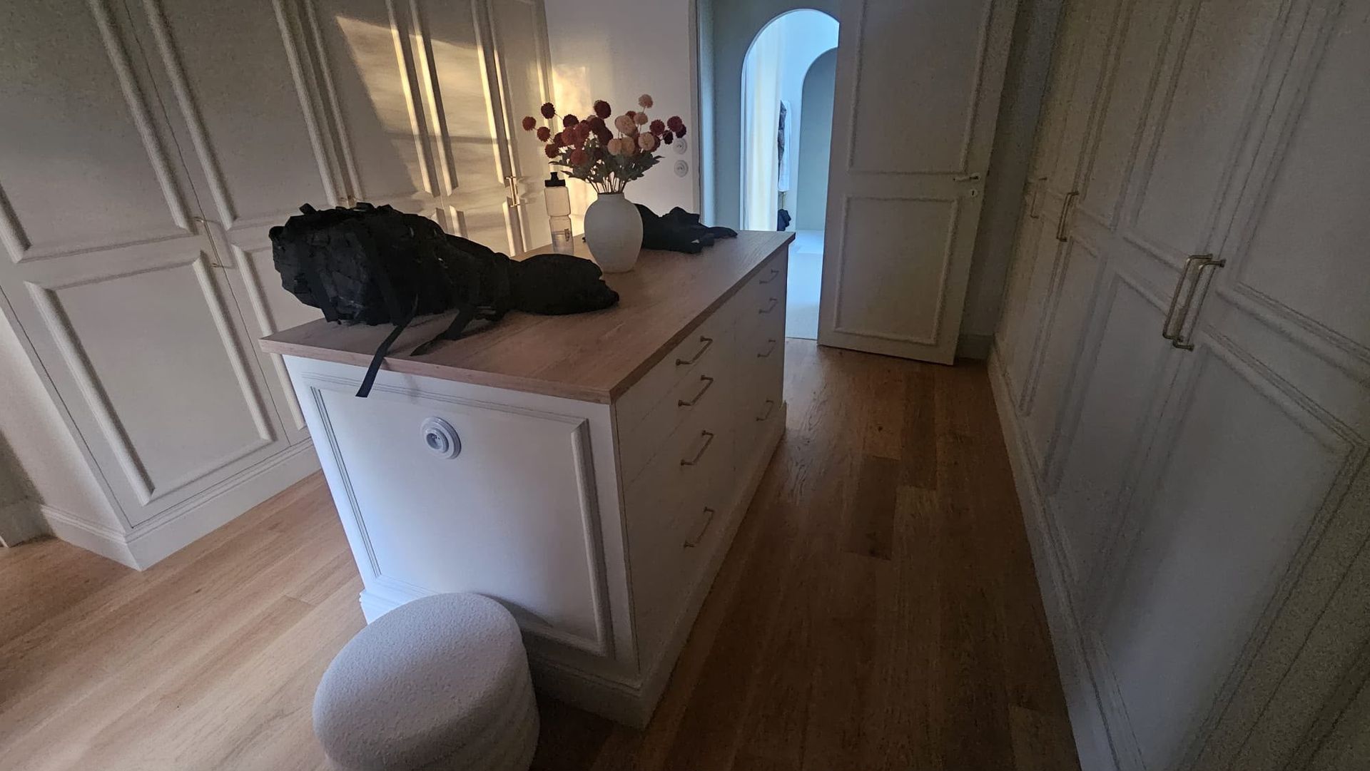Couloir blanc avec une commode, un centre de table en plumes noires, des fleurs et une porte lumineuse au fond.