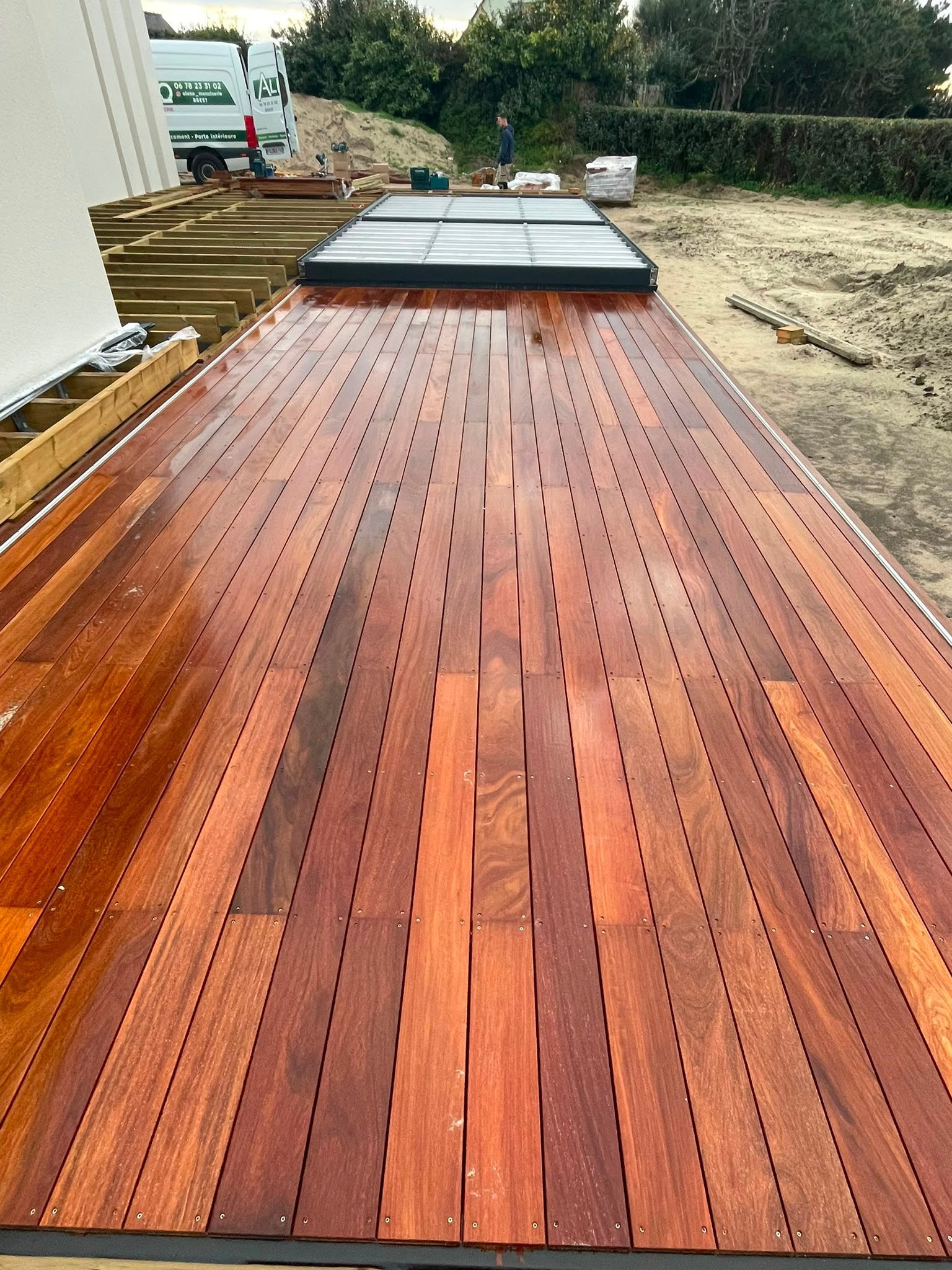 Passerelle en bois menant à une maison moderne, avec des planches rougeâtres et des puits de lumière encadrés de noir devant