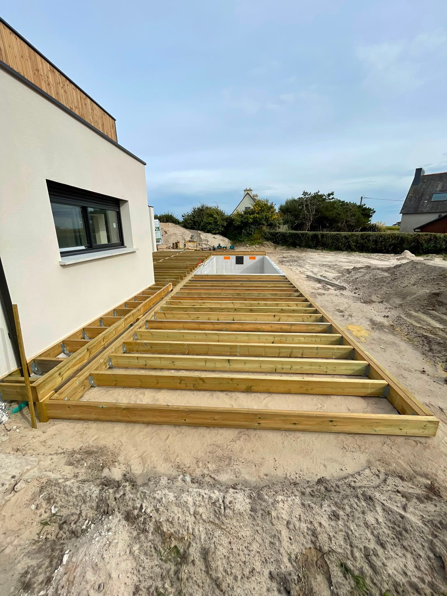 Ossature d’une terrasse en bois à côté d’une maison blanche, sur un terrain sablonneux, en cours de construction