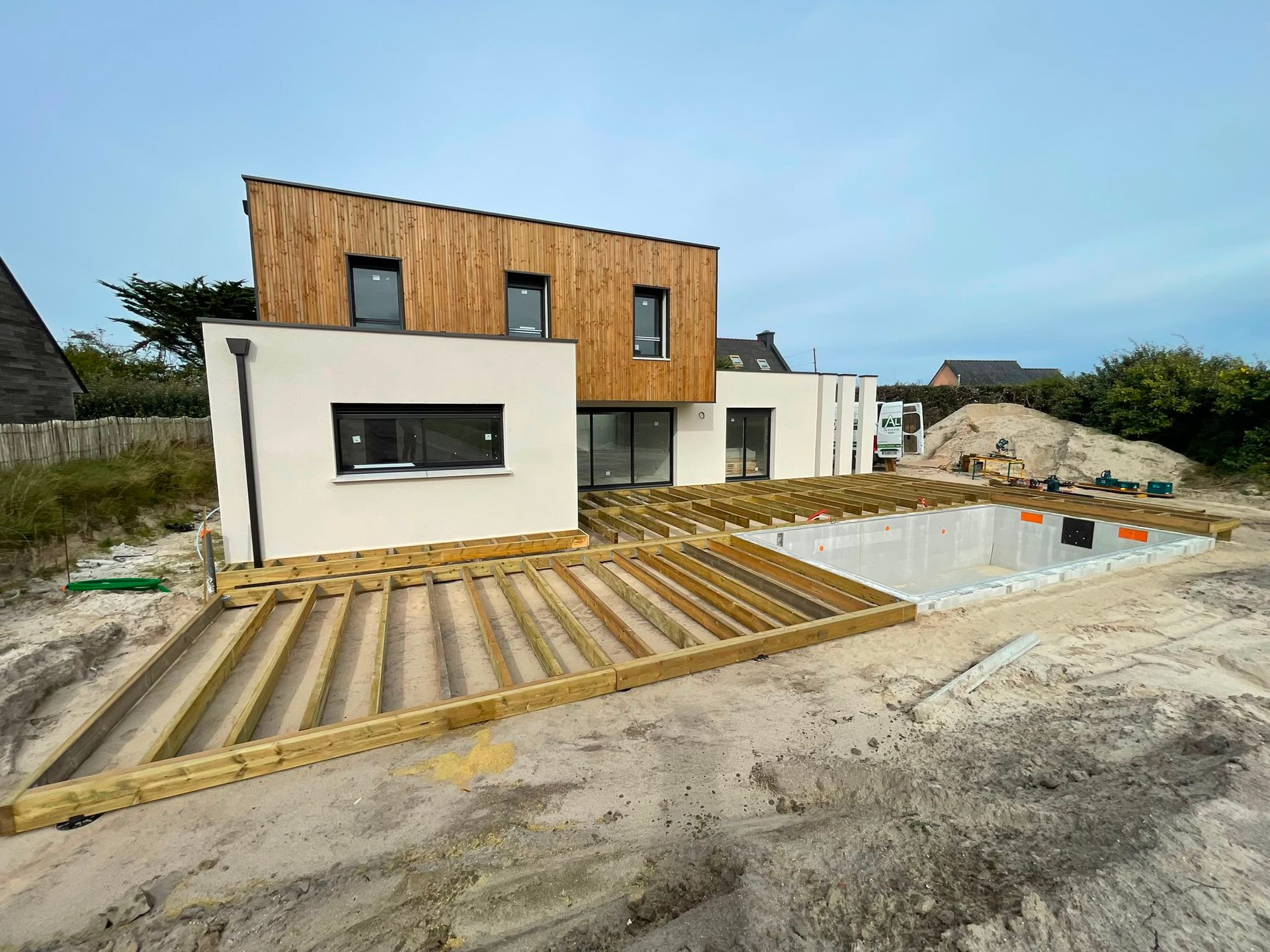 Maison moderne en construction, avec une façade en bois et blanche, au bord d’une piscine et d’un jardin sablonneux