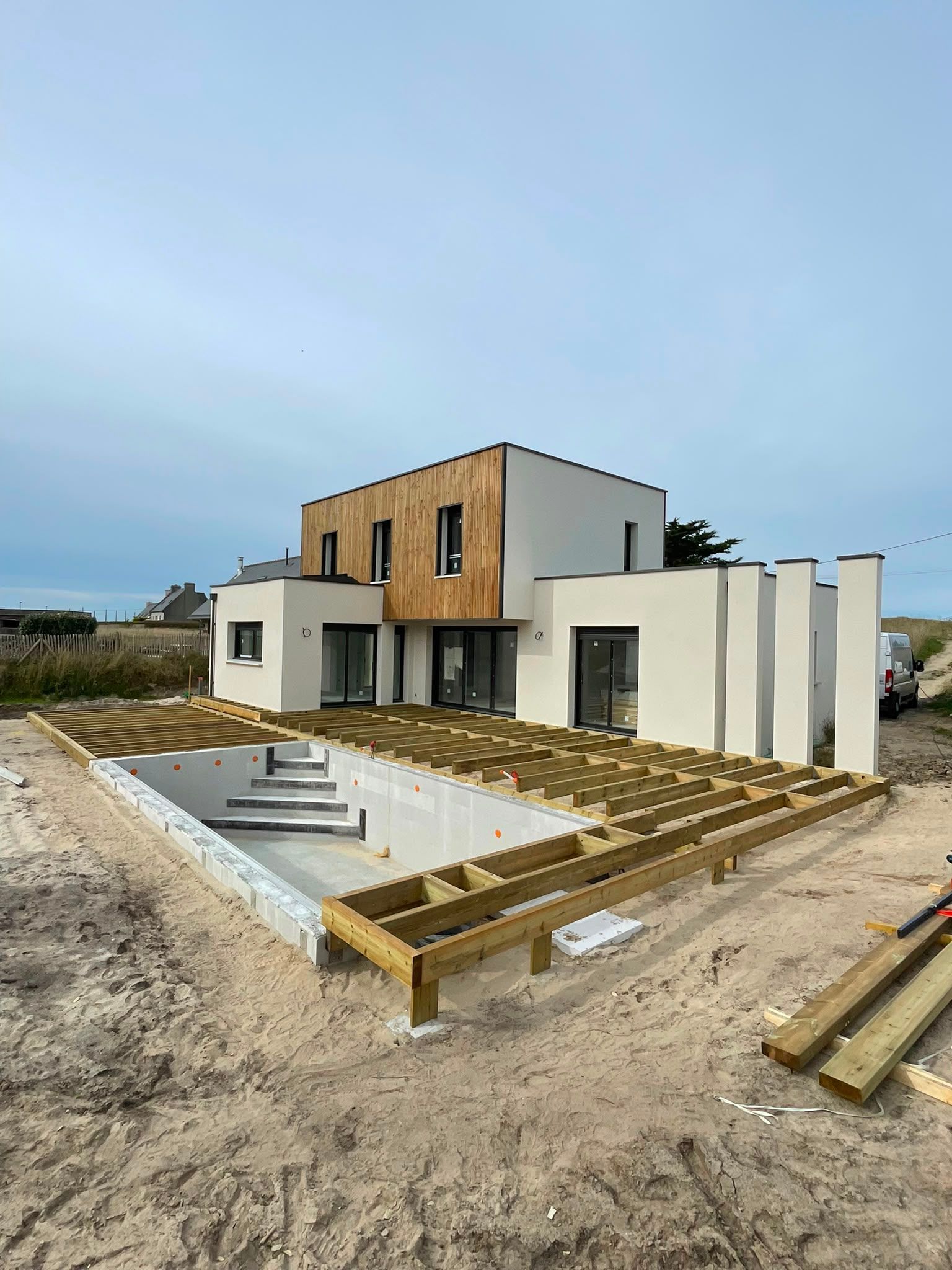 Maison moderne en construction, avec des murs blancs, un bardage en bois et une cour recouverte de sable avec une piscine.