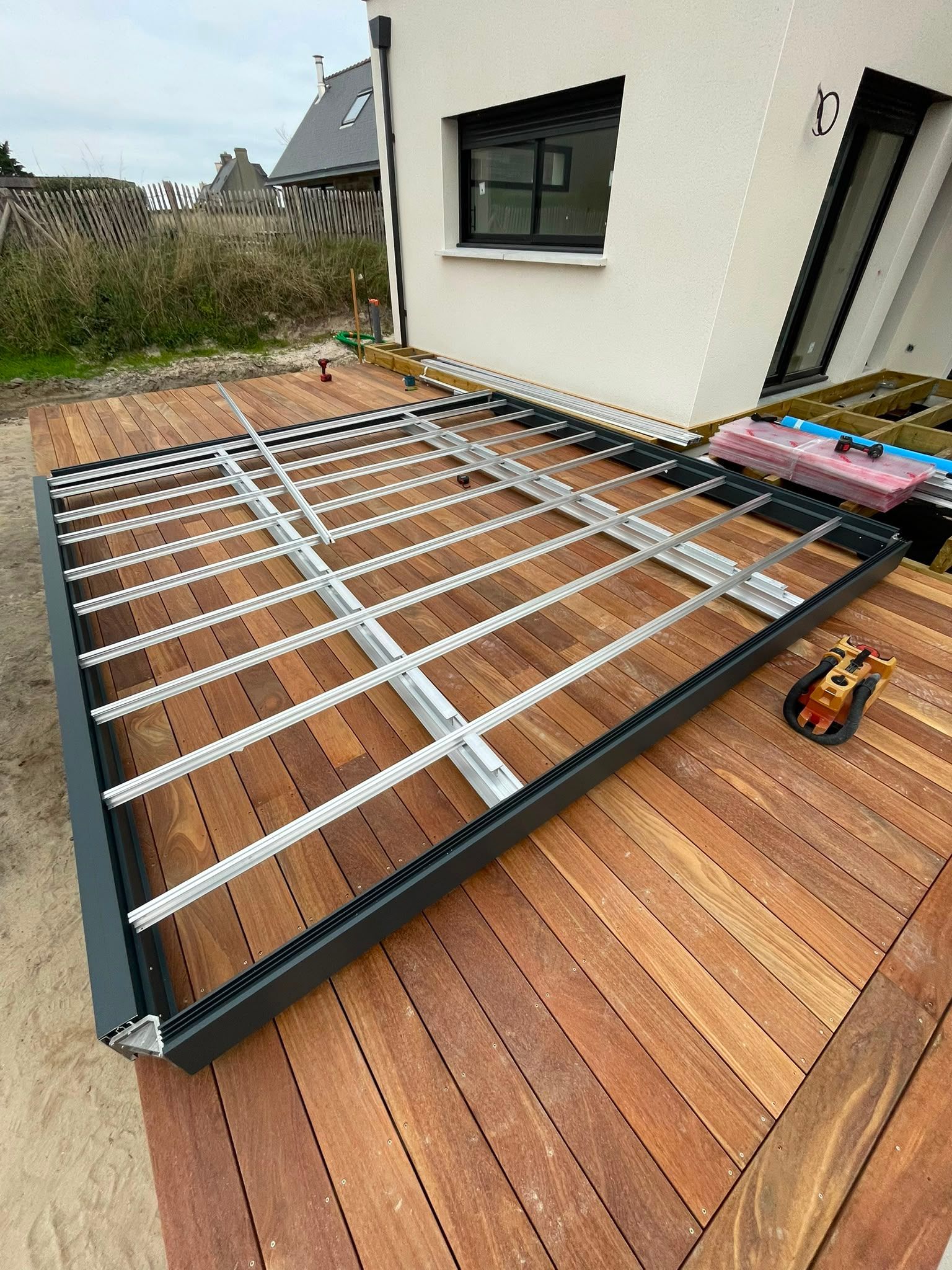 Cadre en métal noir sur une terrasse en bois, à côté d’une maison, avec des lattes blanches en cours d’installation.