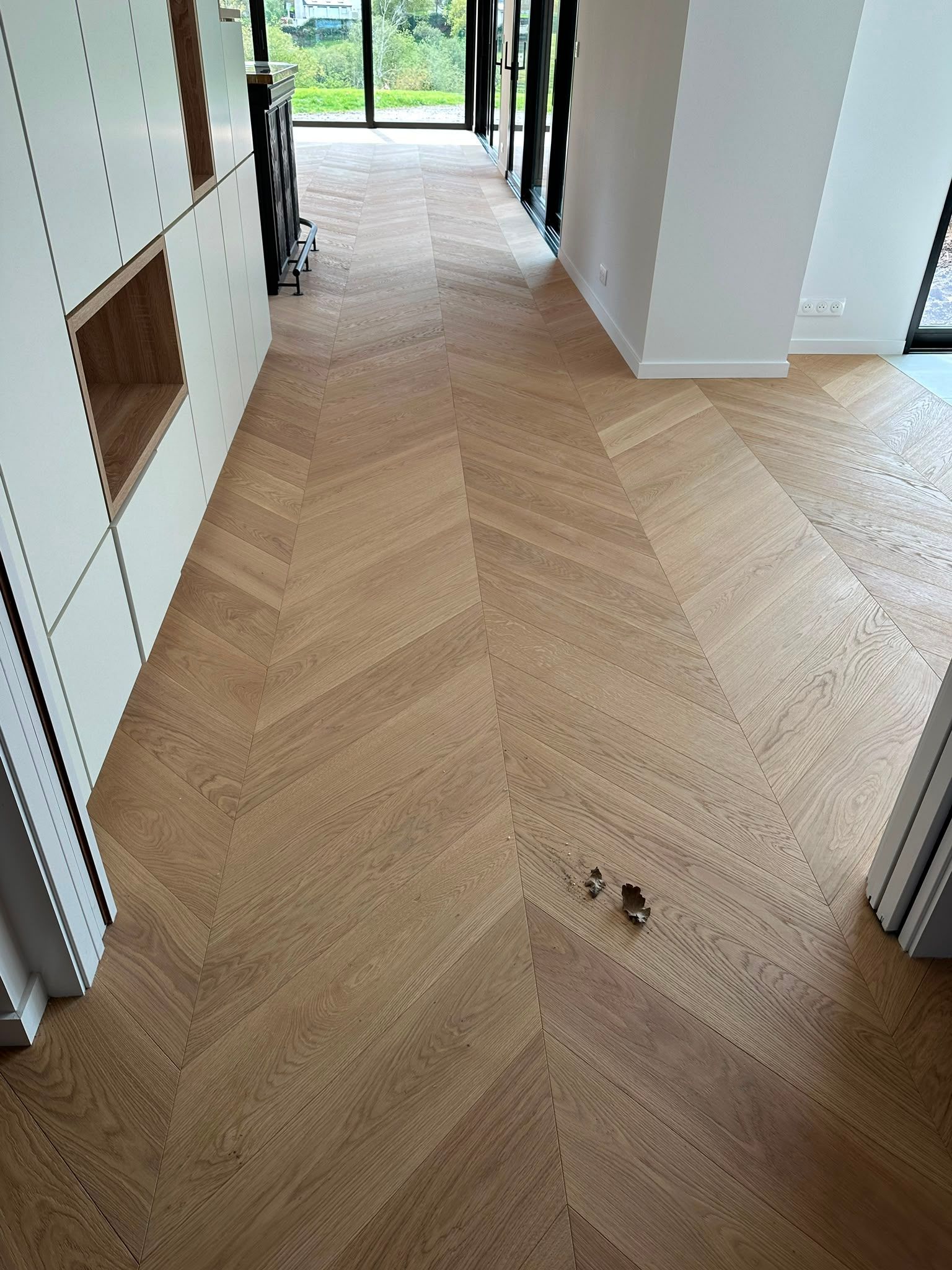 Couloir avec un parquet en bois clair à chevrons, des murs blancs et une porte donnant sur une vue extérieure lumineuse