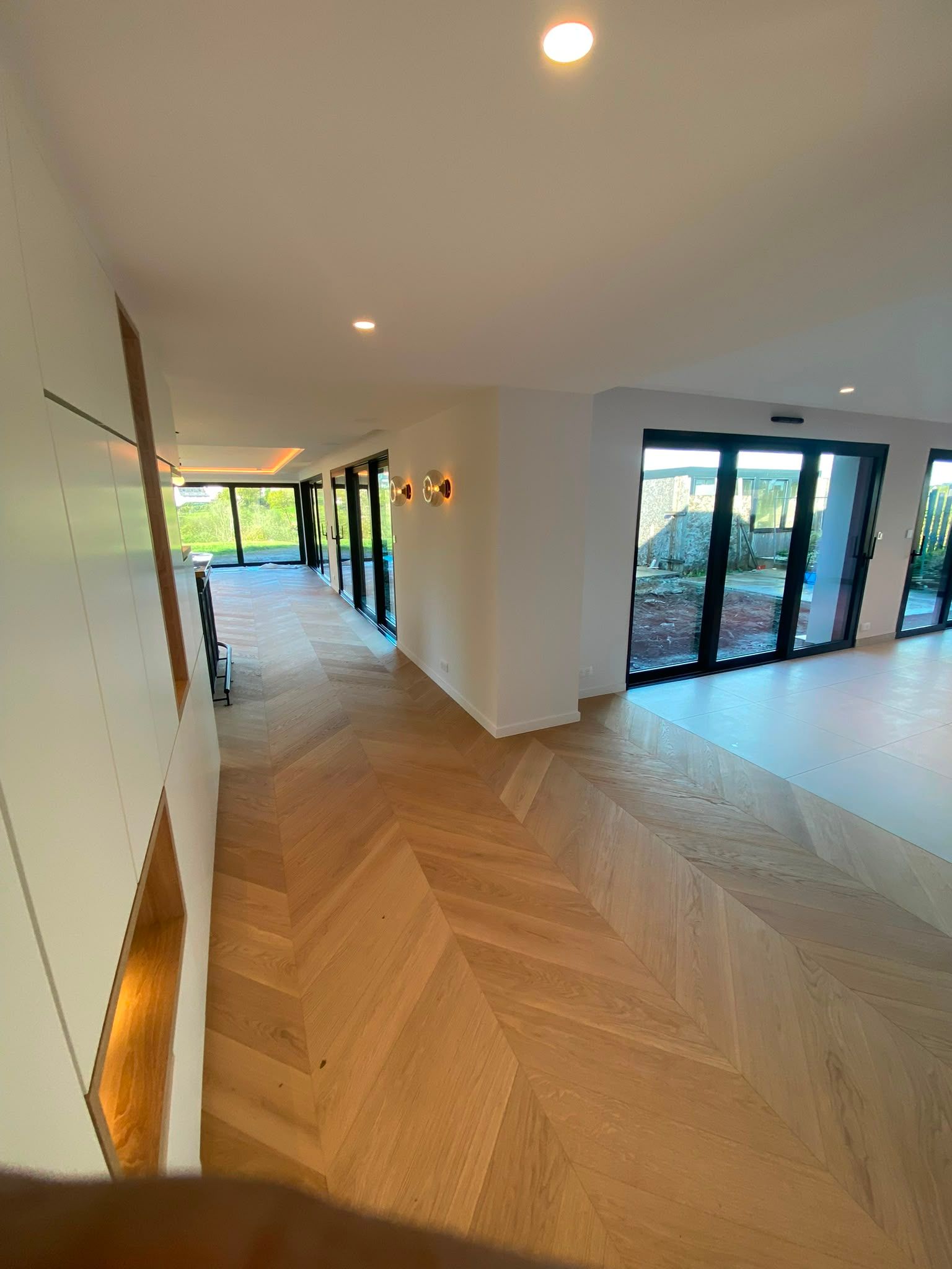 Couloir moderne avec parquet en chevrons, murs blancs et grandes portes vitrées donnant sur une terrasse lumineuse