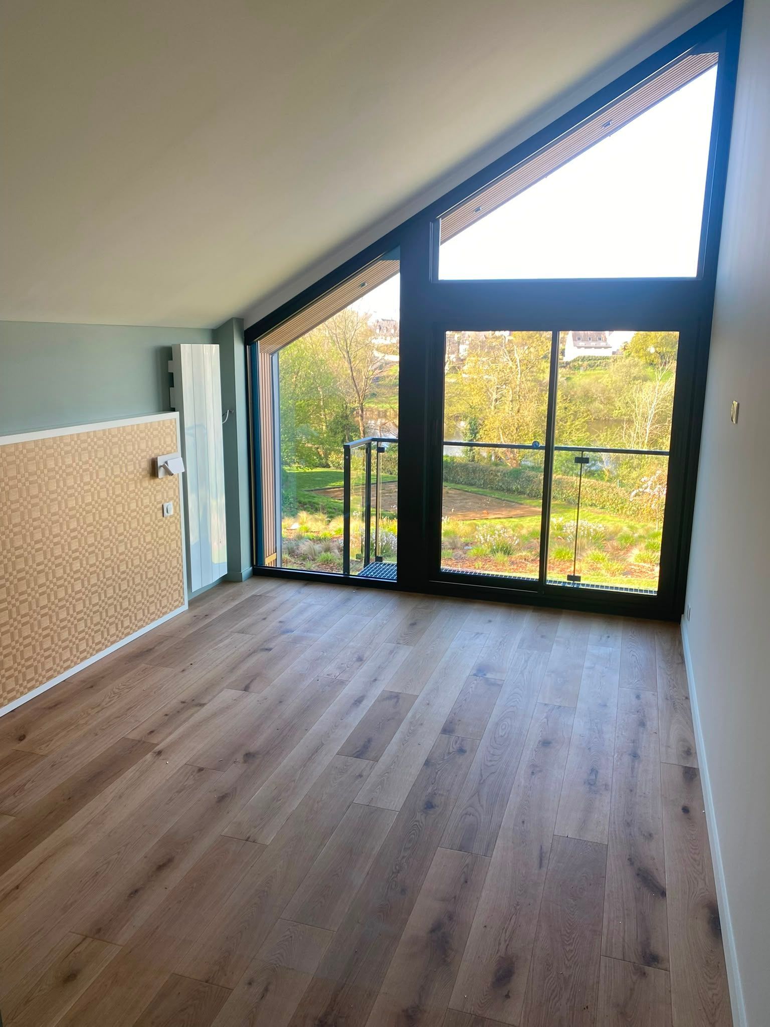 Pièce mansardée vide avec un plafond en pente, de grandes fenêtres à cadres noirs et un parquet donnant sur des arbres.