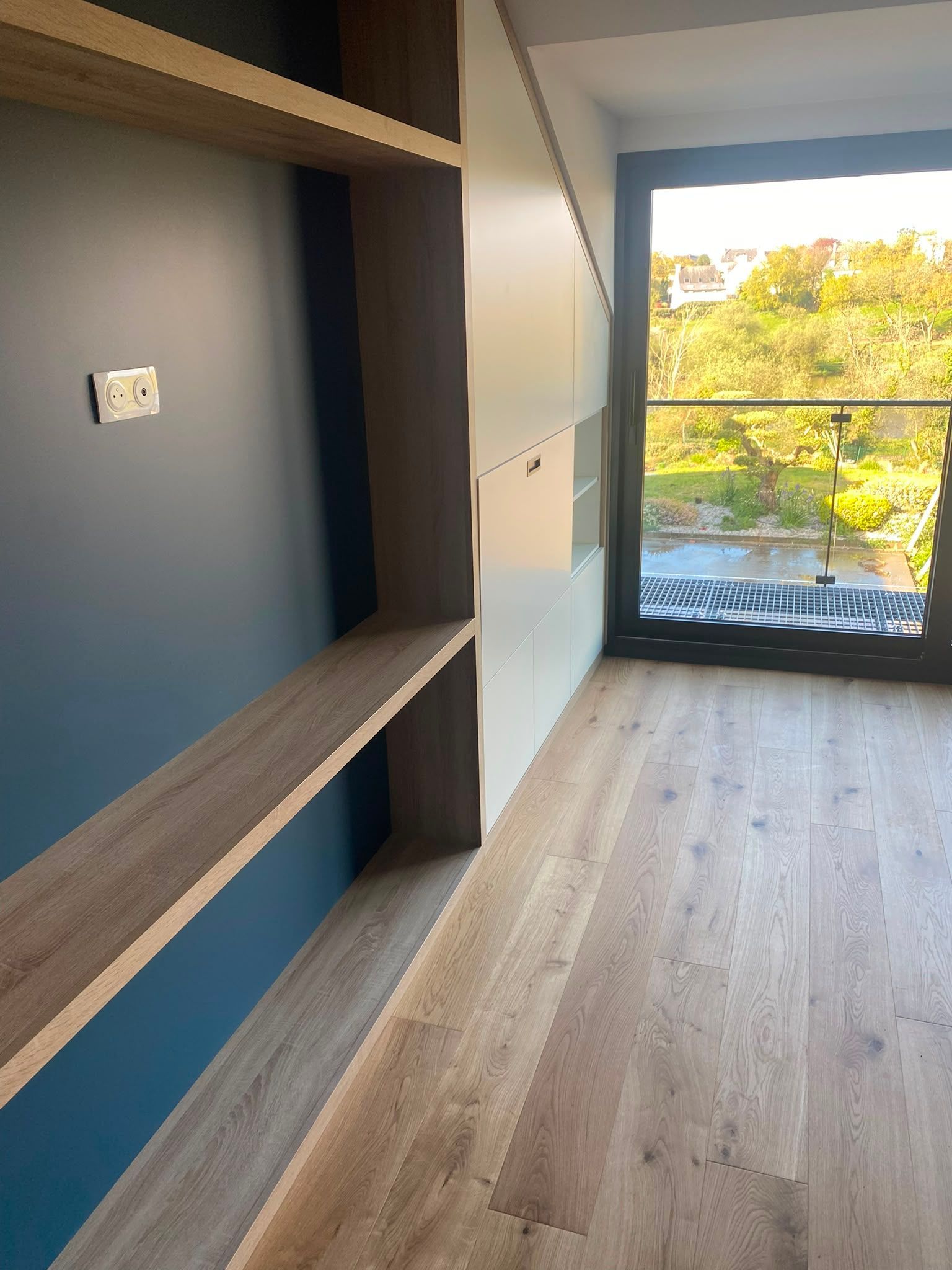Pièce moderne vide avec des étagères intégrées, un mur d’accent bleu, un parquet et une grande fenêtre donnant sur des arbres