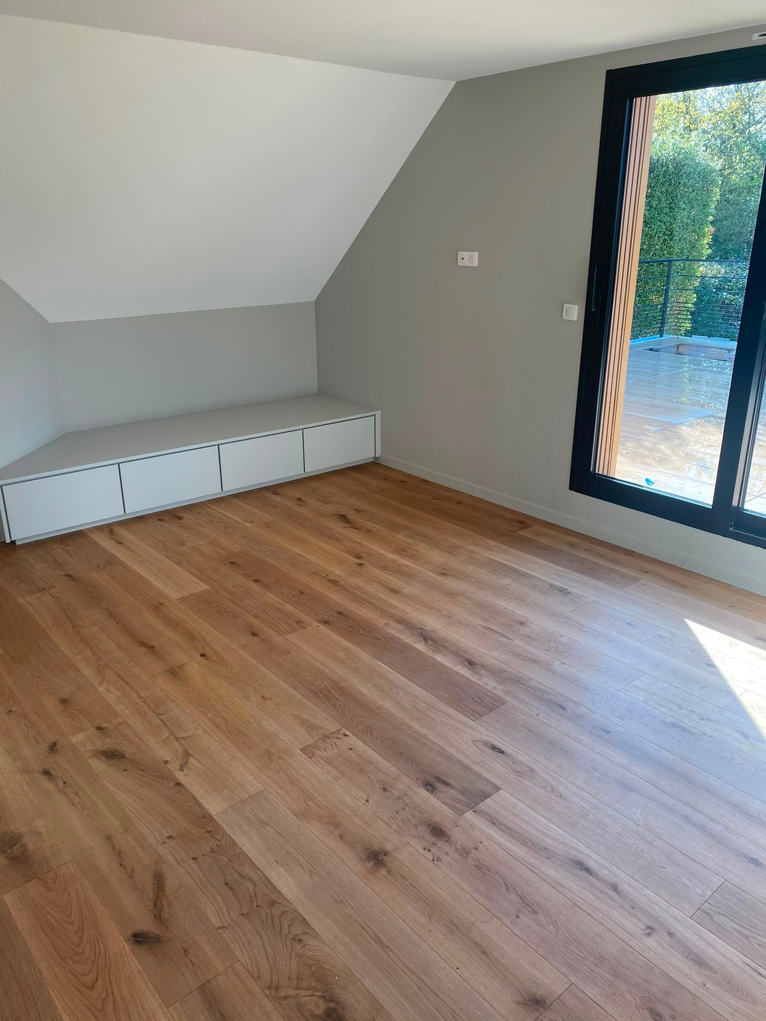 Pièce vide avec parquet, murs blancs en pente, rangements intégrés et grande porte vitrée donnant sur un jardin.