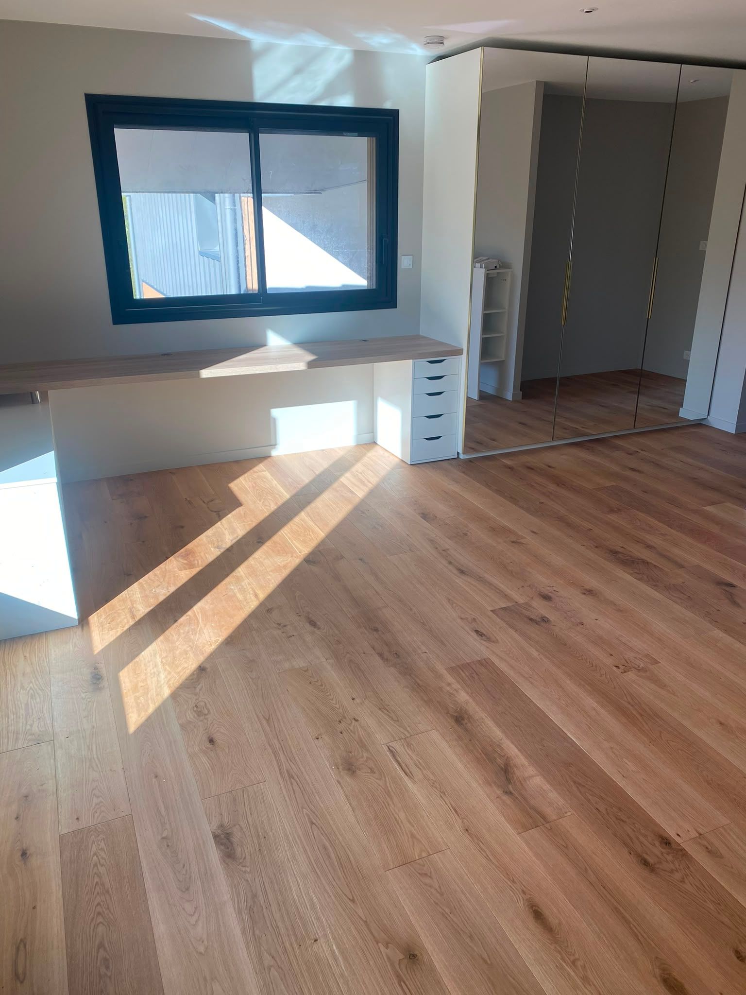 Pièce vide avec parquet, fenêtre, bureau intégré et portes de placard ouvertes.