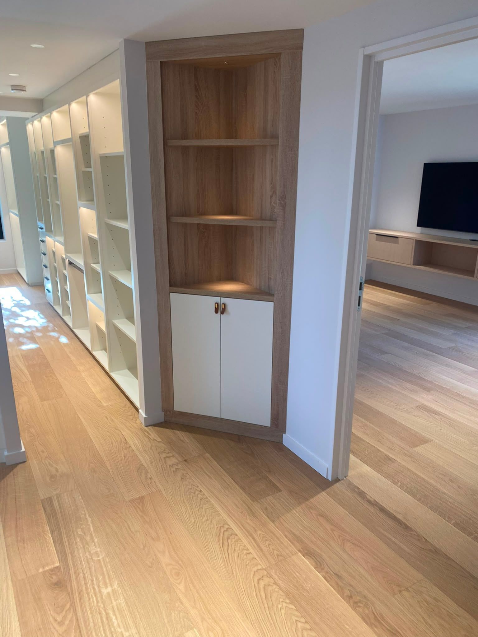 Couloir avec parquet, étagères intégrées et porte donnant sur une pièce avec une télévision et un meuble.
