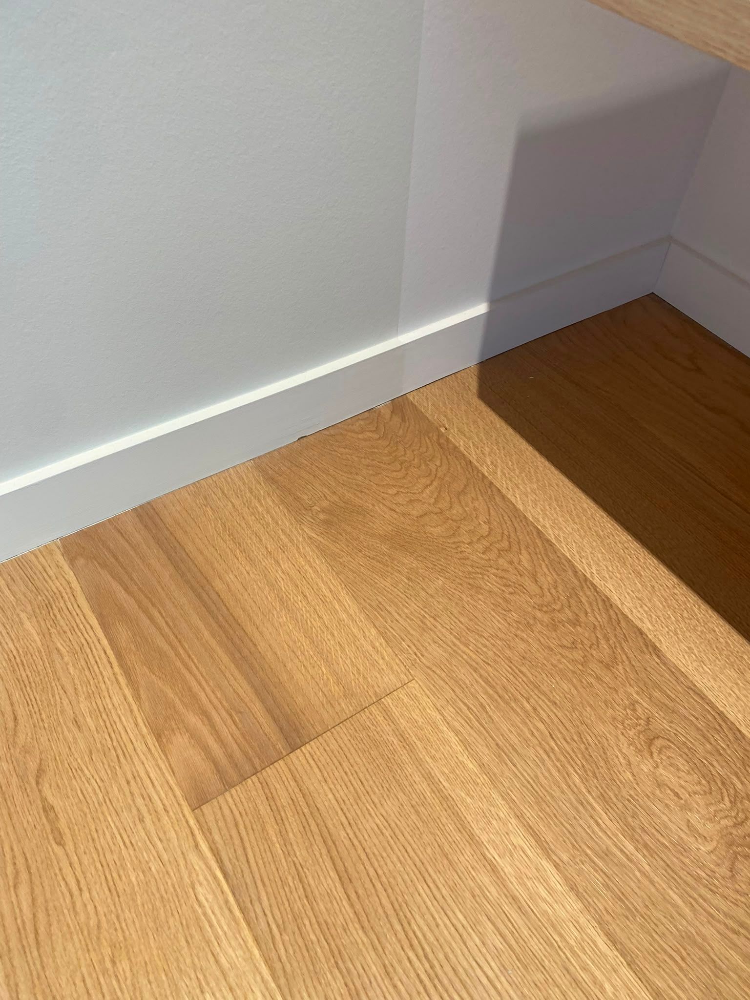 Coin d’une pièce avec un parquet clair, une plinthe blanche et une zone de sol plus sombre près d’une ouverture dans le mur