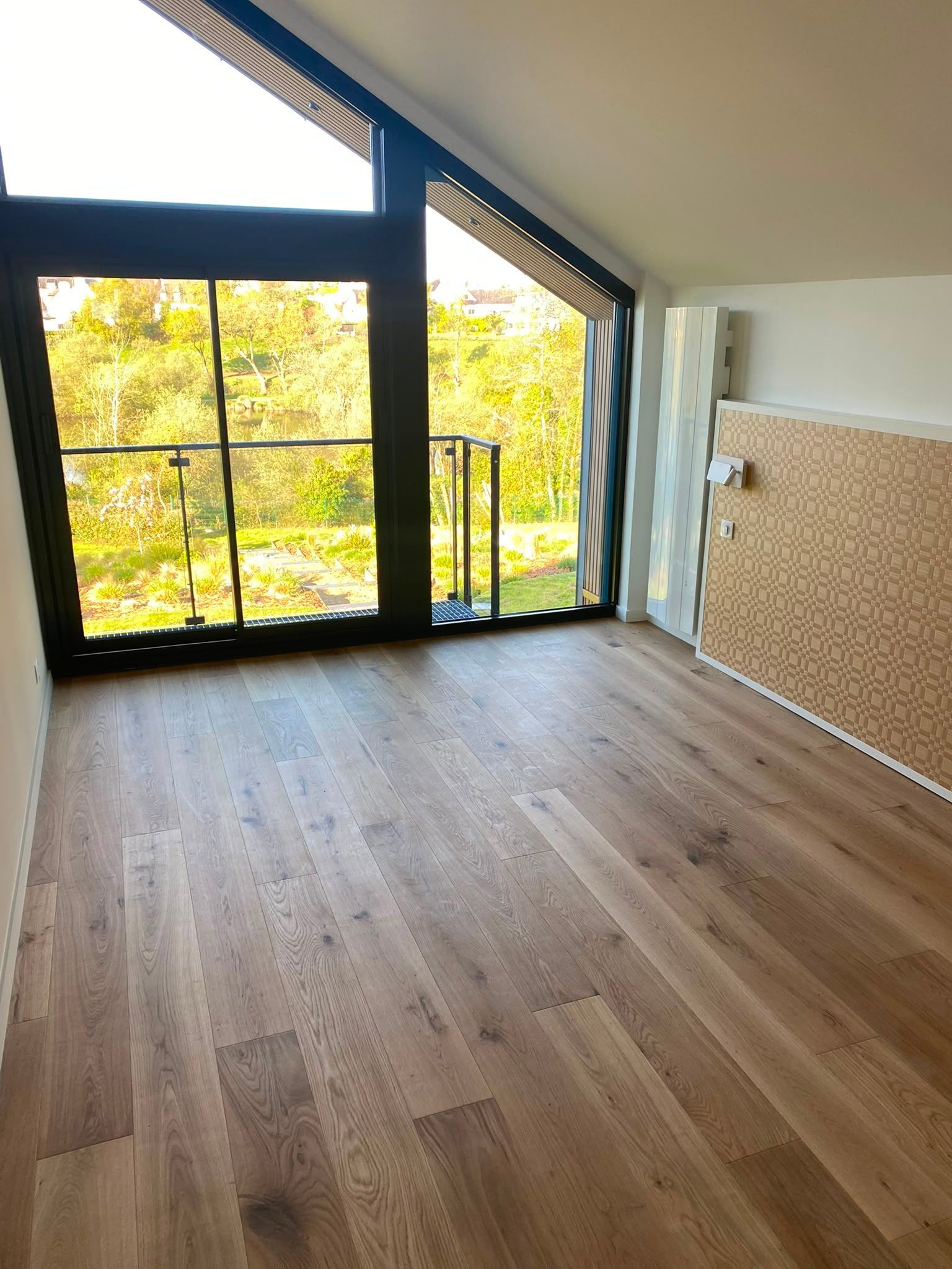 Pièce mansardée vide avec un parquet, un plafond en pente, de grandes fenêtres à cadre noir et un mur d’accent en briques.