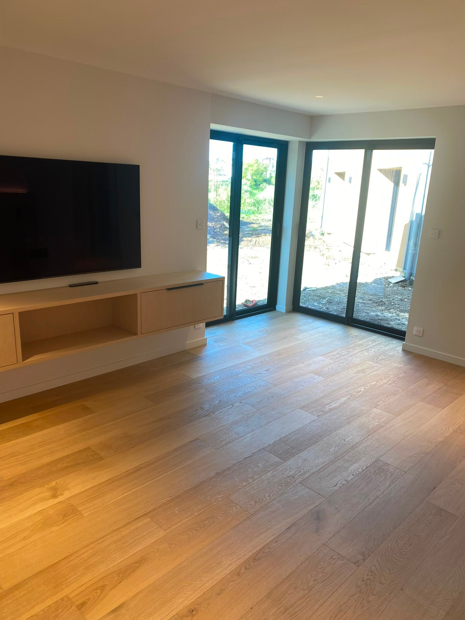 Salon lumineux avec parquet, télévision murale et portes vitrées ouvertes sur une terrasse et un jardin.