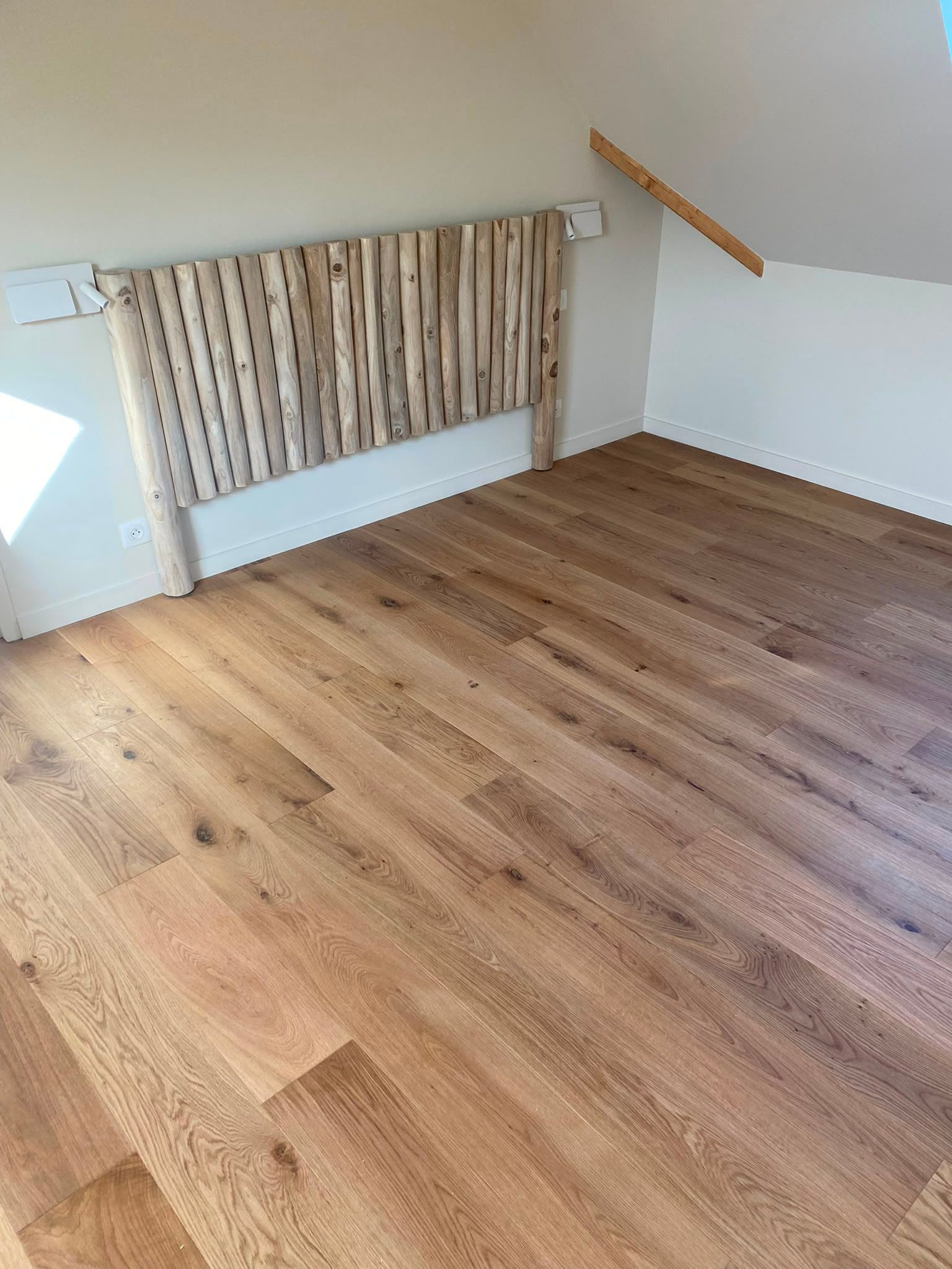 Pièce vide avec un parquet, un plafond en pente, un radiateur et un rideau à motifs devant une fenêtre.