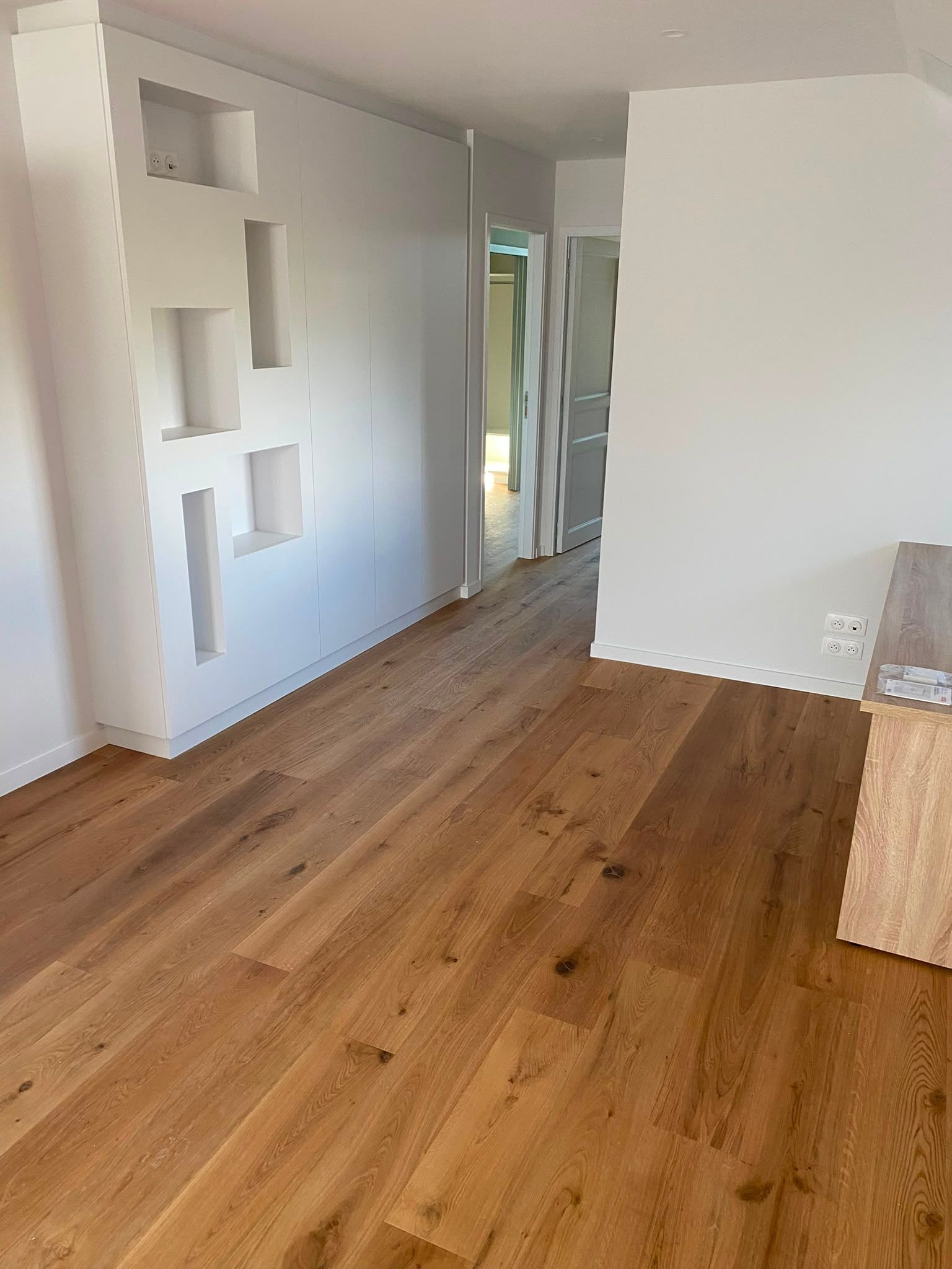 Pièce moderne vide avec parquet, murs blancs et niches murales intégrées, donnant sur un couloir.