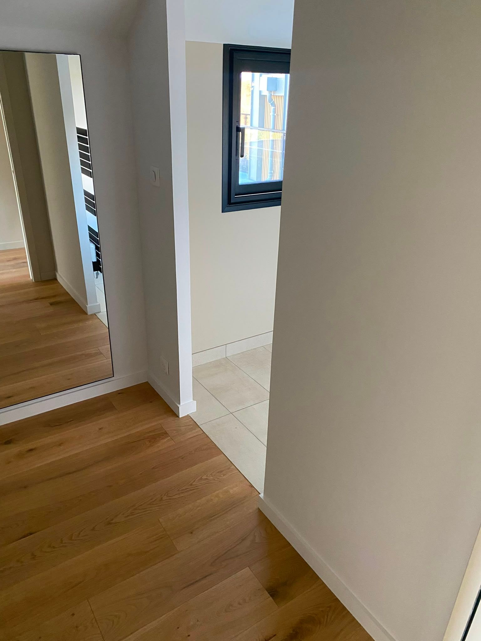 Couloir lumineux avec parquet, murs blancs et petite fenêtre à côté d’une zone carrelée