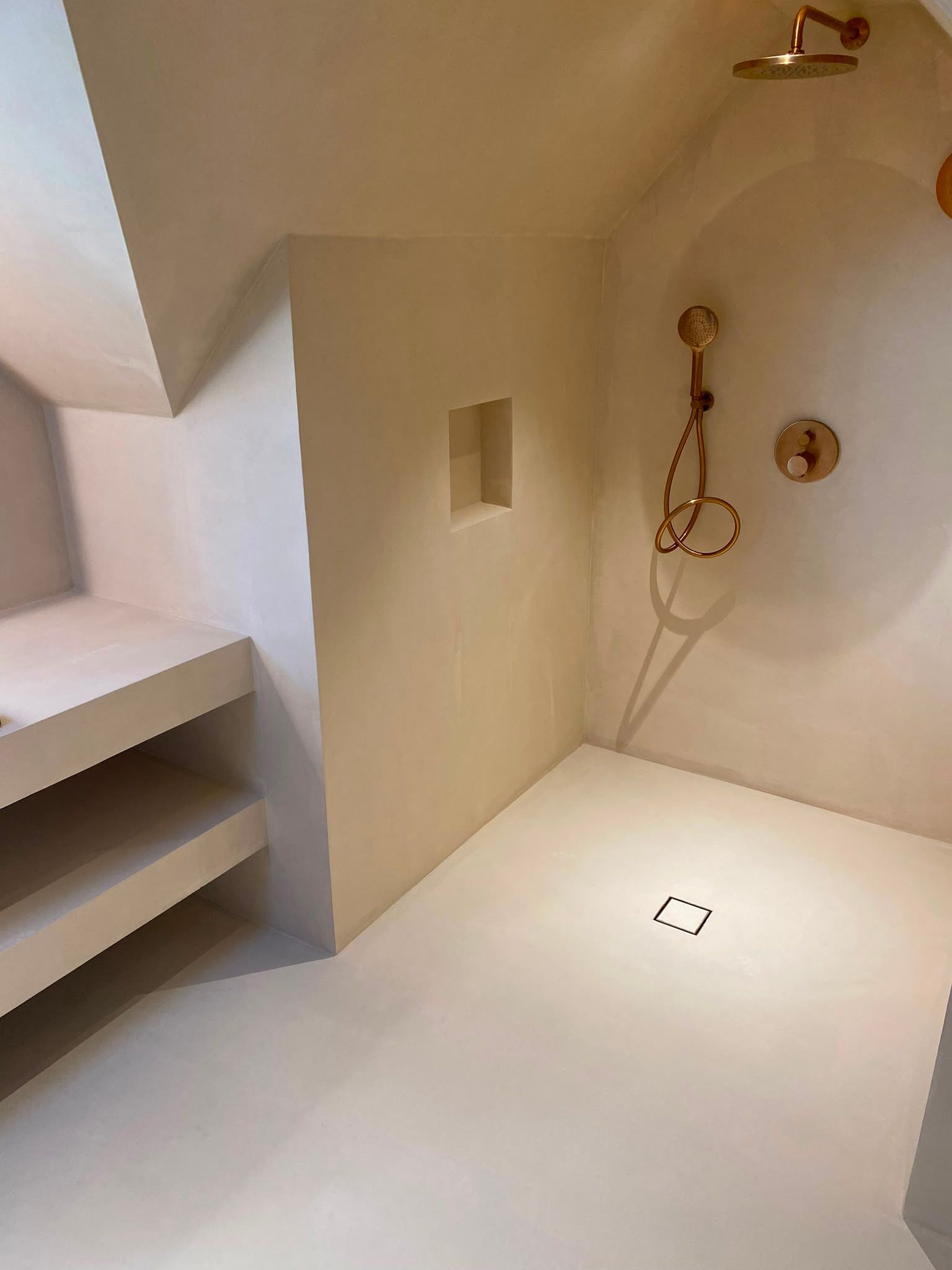 Petite cabine de douche blanche avec étagères intégrées, douchette à main et siphon de sol.