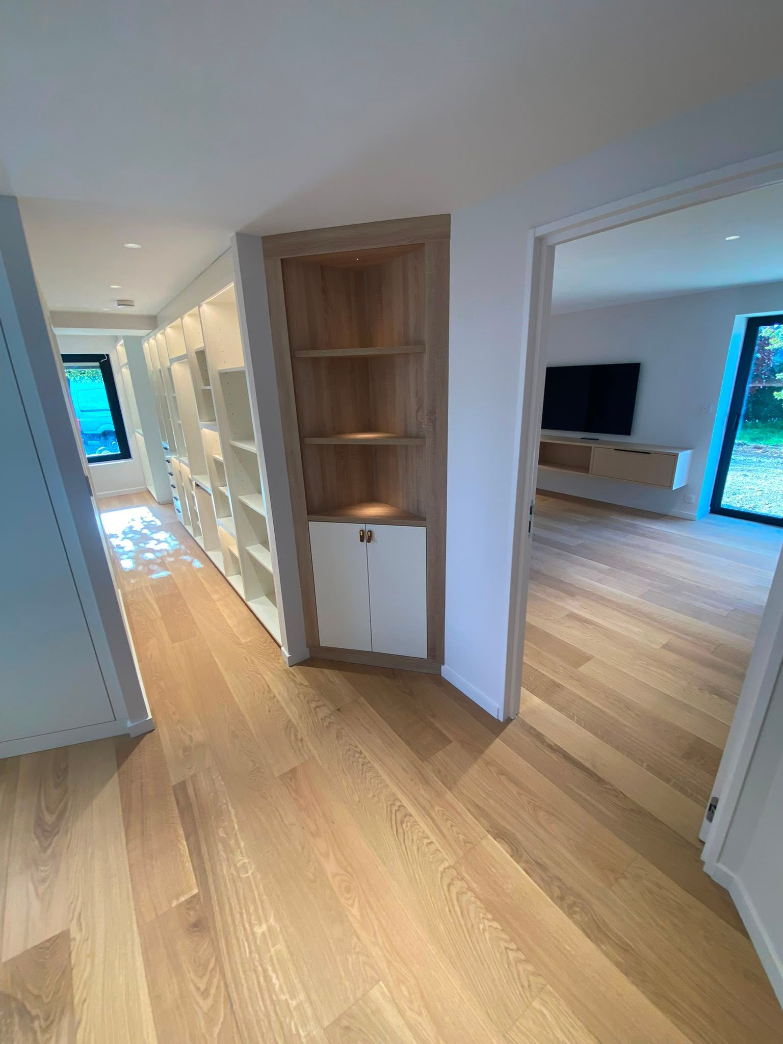Couloir lumineux avec parquet clair, étagères intégrées et salon attenant visible à travers une porte ouverte.