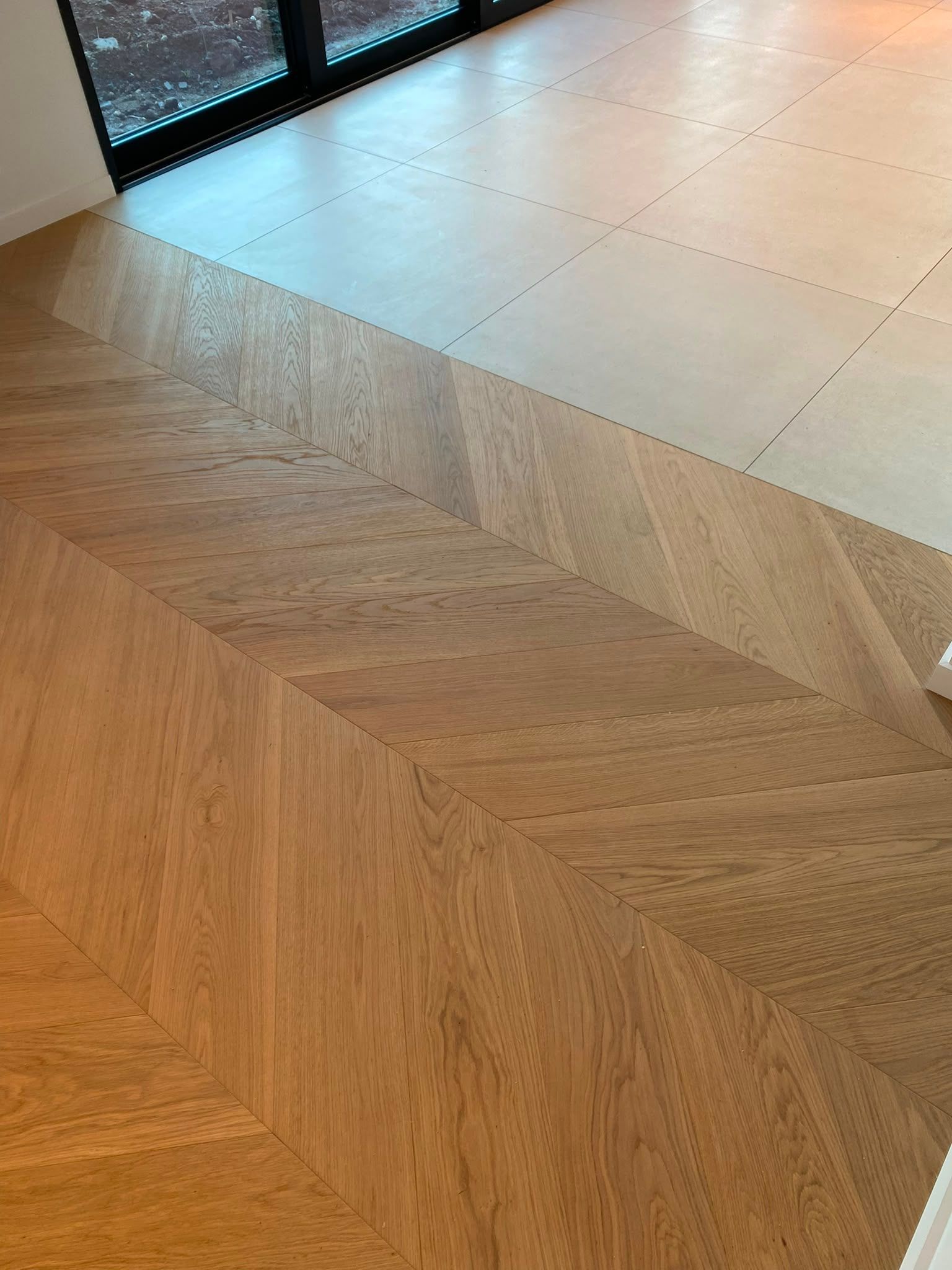 Parquet clair en chevrons qui rejoint un carrelage carré pâle près d’une porte vitrée.