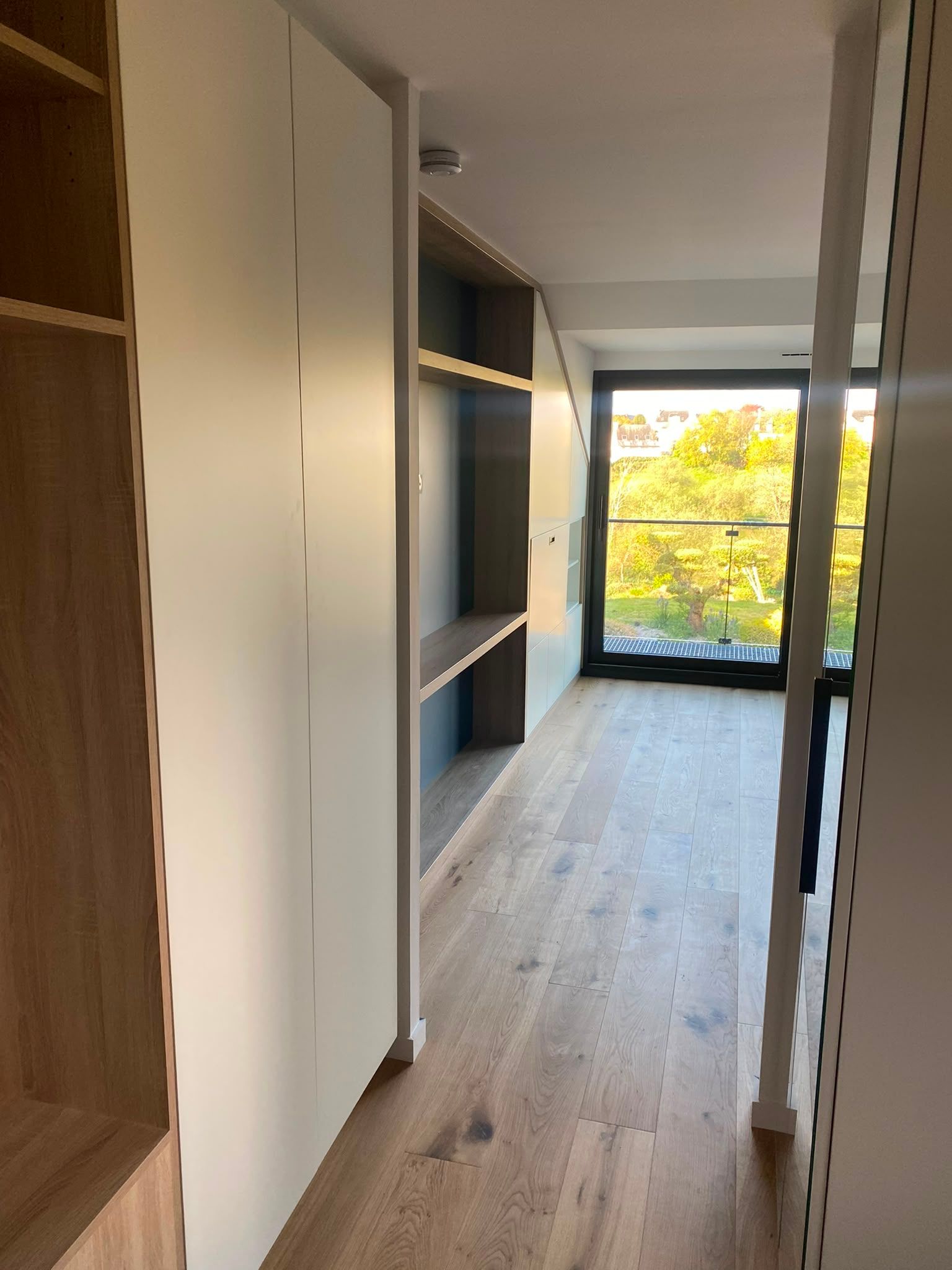 Couloir étroit avec des étagères intégrées, un parquet clair et une grande fenêtre donnant sur des arbres.