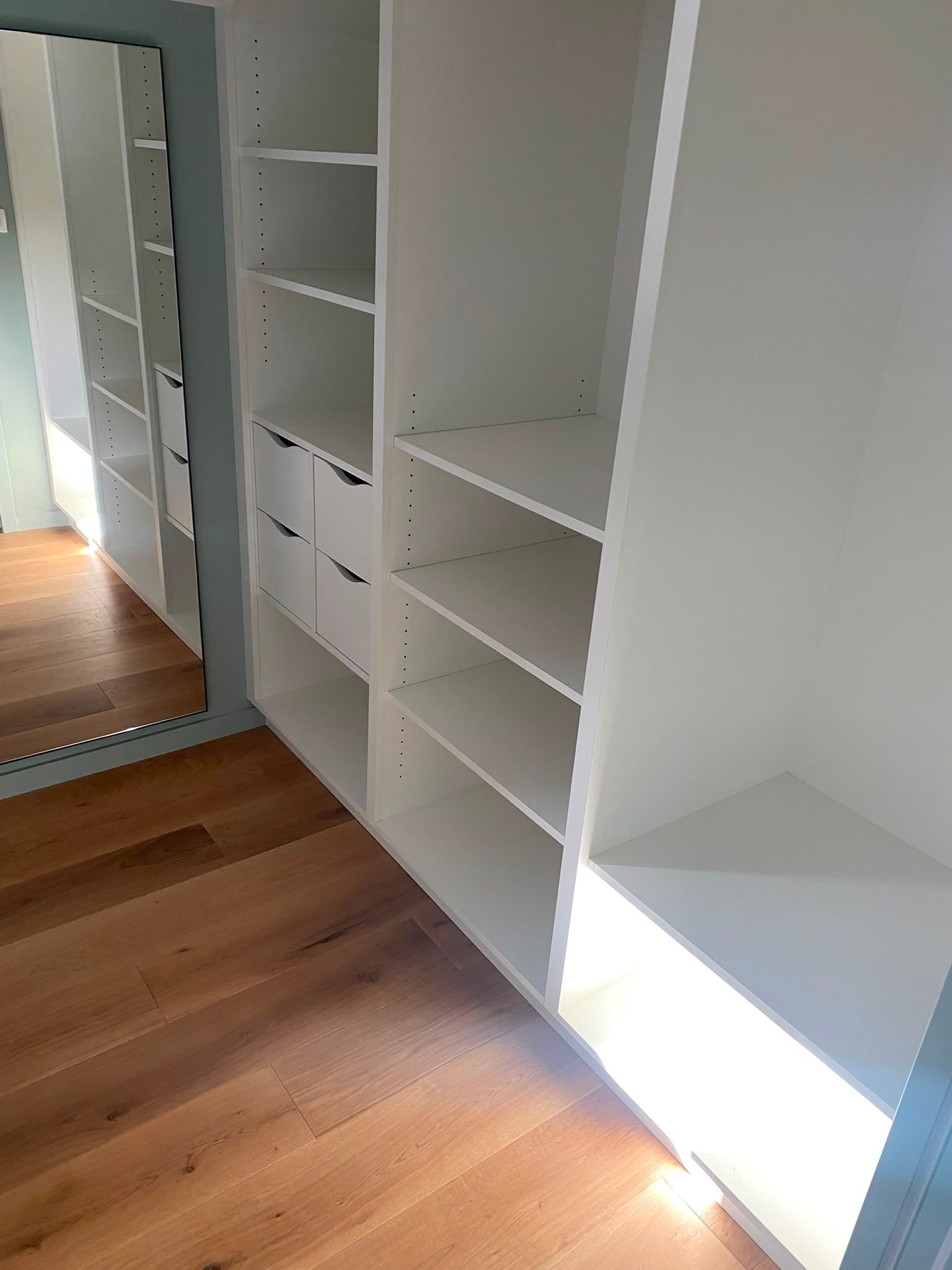 Étagères de placard blanches intégrées à côté d’une porte miroir, avec le soleil qui éclaire le parquet.