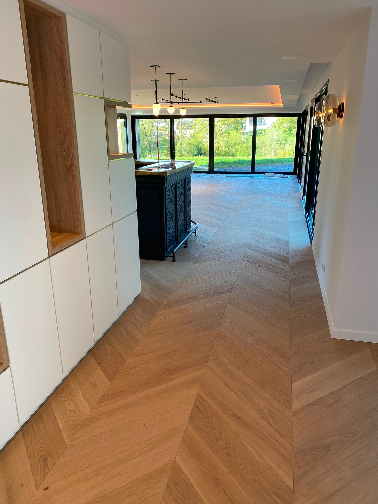 Cuisine et séjour modernes avec parquet en chevrons, placards blancs et grandes fenêtres donnant sur la verdure