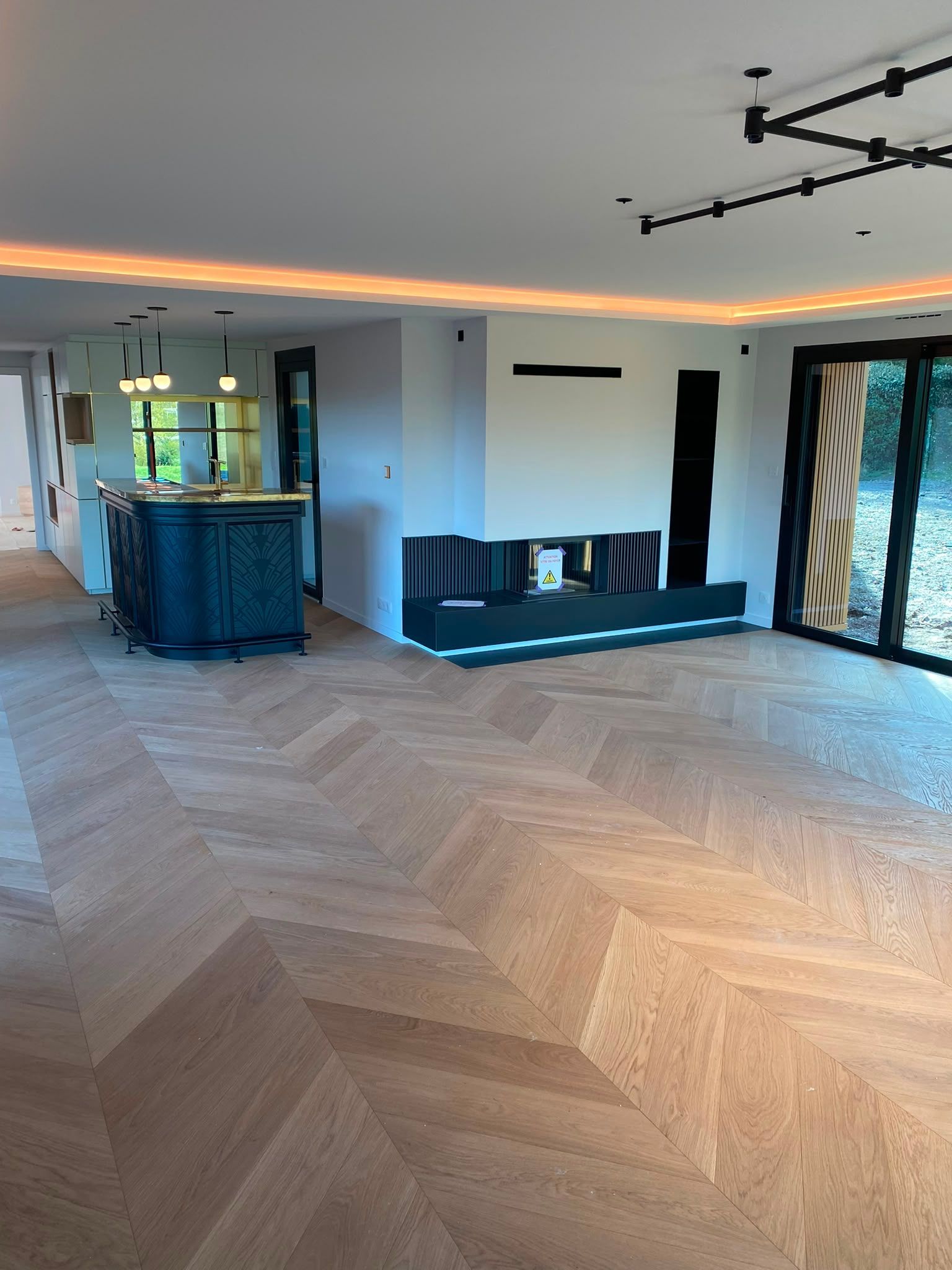 Salon moderne avec parquet en chevrons, bar bleu, canapé noir et portes-fenêtres coulissantes avec rideaux