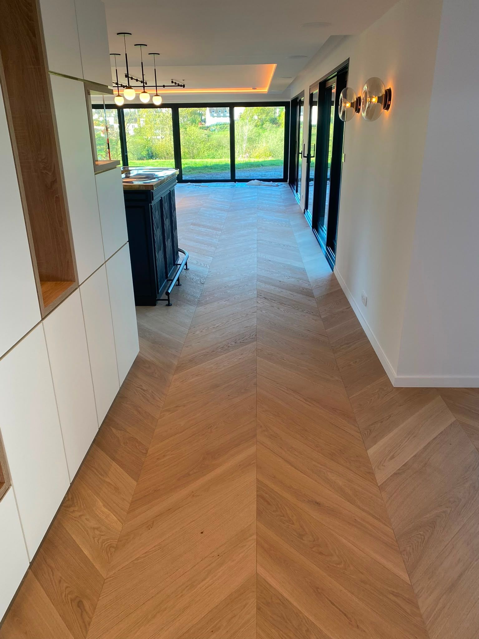 Hall d’entrée moderne avec parquet en chevrons, appliques murales et grandes fenêtres donnant sur la verdure.