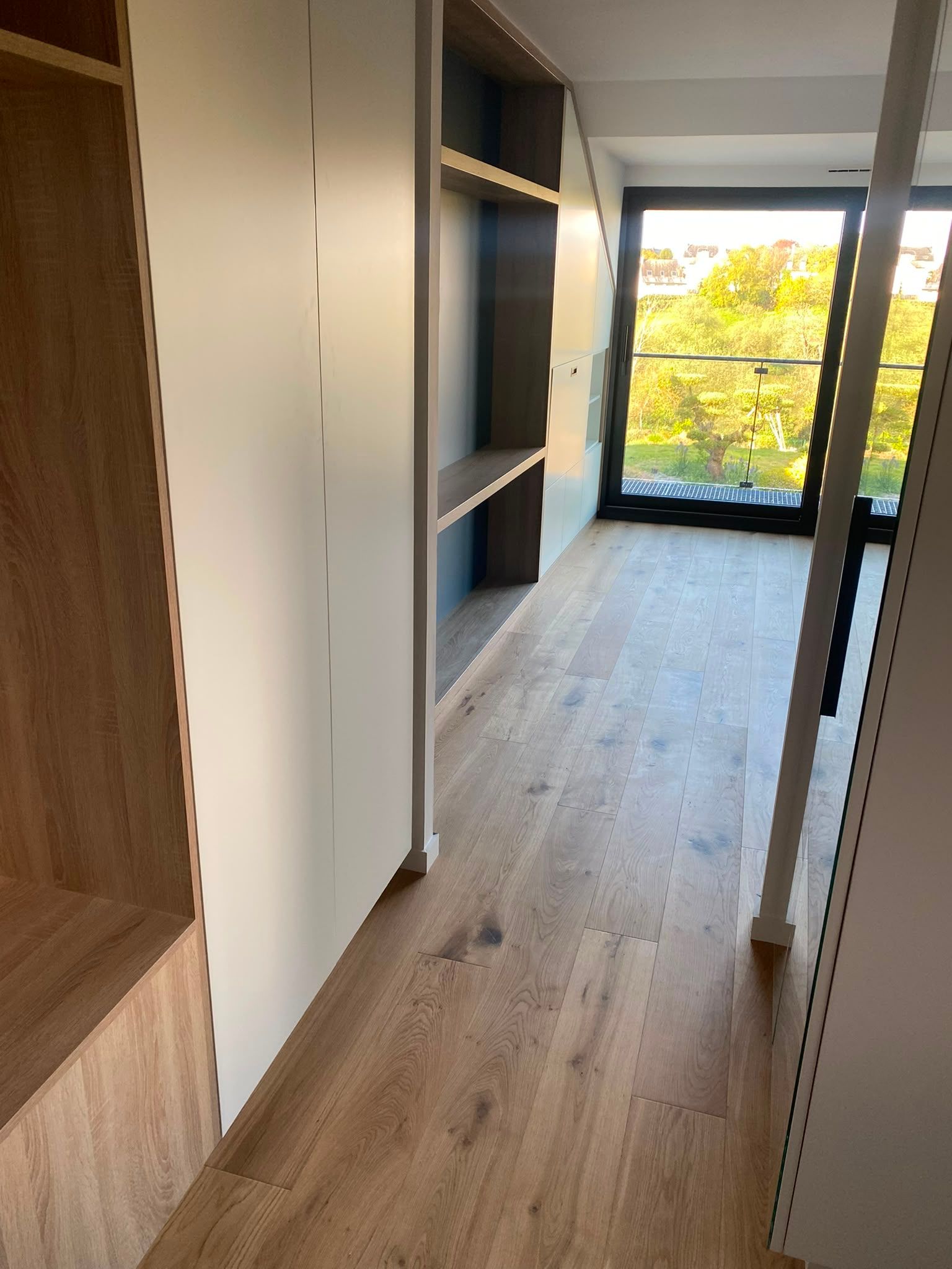 Couloir étroit avec parquet, étagères intégrées et grande fenêtre donnant sur les arbres.