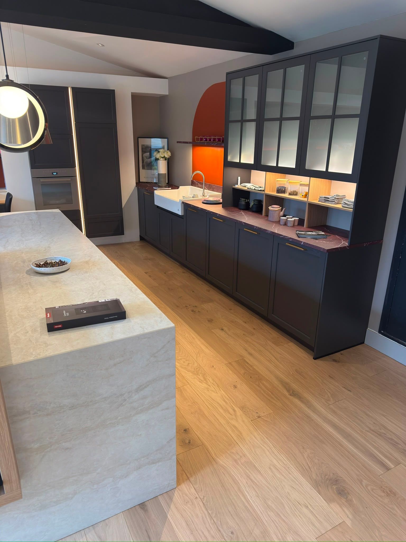 Cuisine moderne avec un grand îlot, des armoires foncées, un plancher en bois et un mur de placards vitrés.