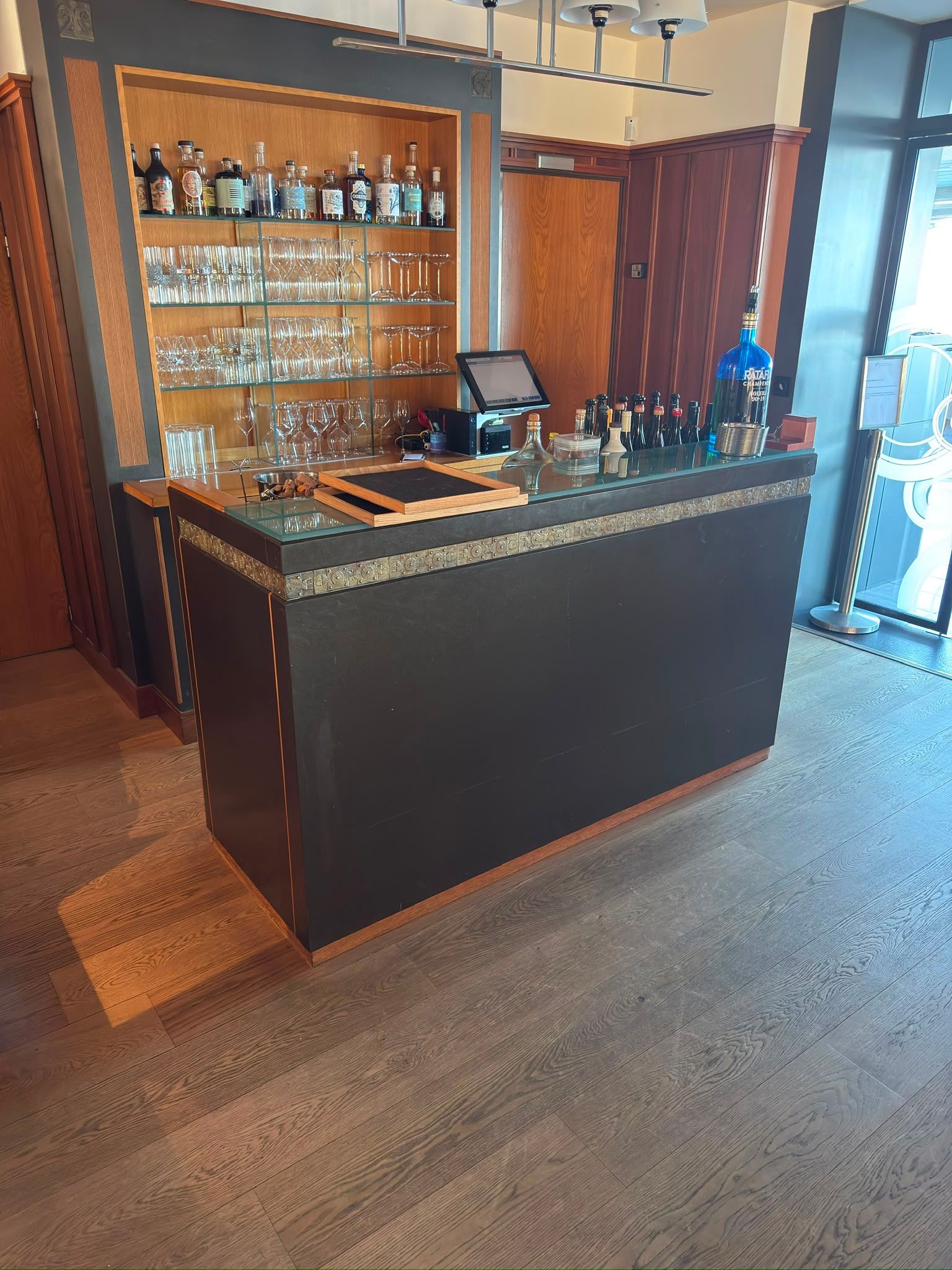 Bar d’hôtel avec comptoir en bois sombre, étagères en verre et sièges dans un salon chaleureux et moderne.