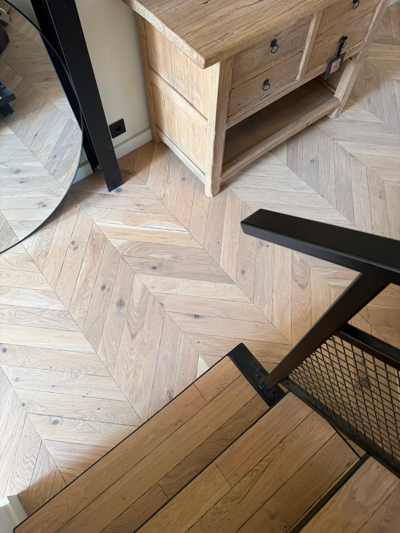 Parquet en chevrons, fauteuil en osier, pied de table noir et meuble tressé dans un intérieur chaleureux.