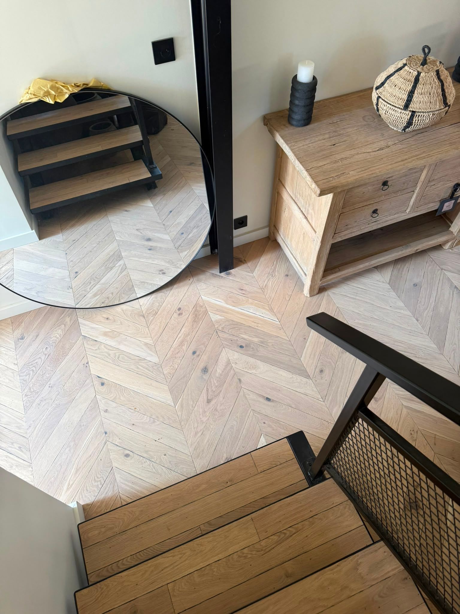 Entrée avec parquet à chevrons, miroir rond, console en bois et escalier avec un panier tressé sur le palier
